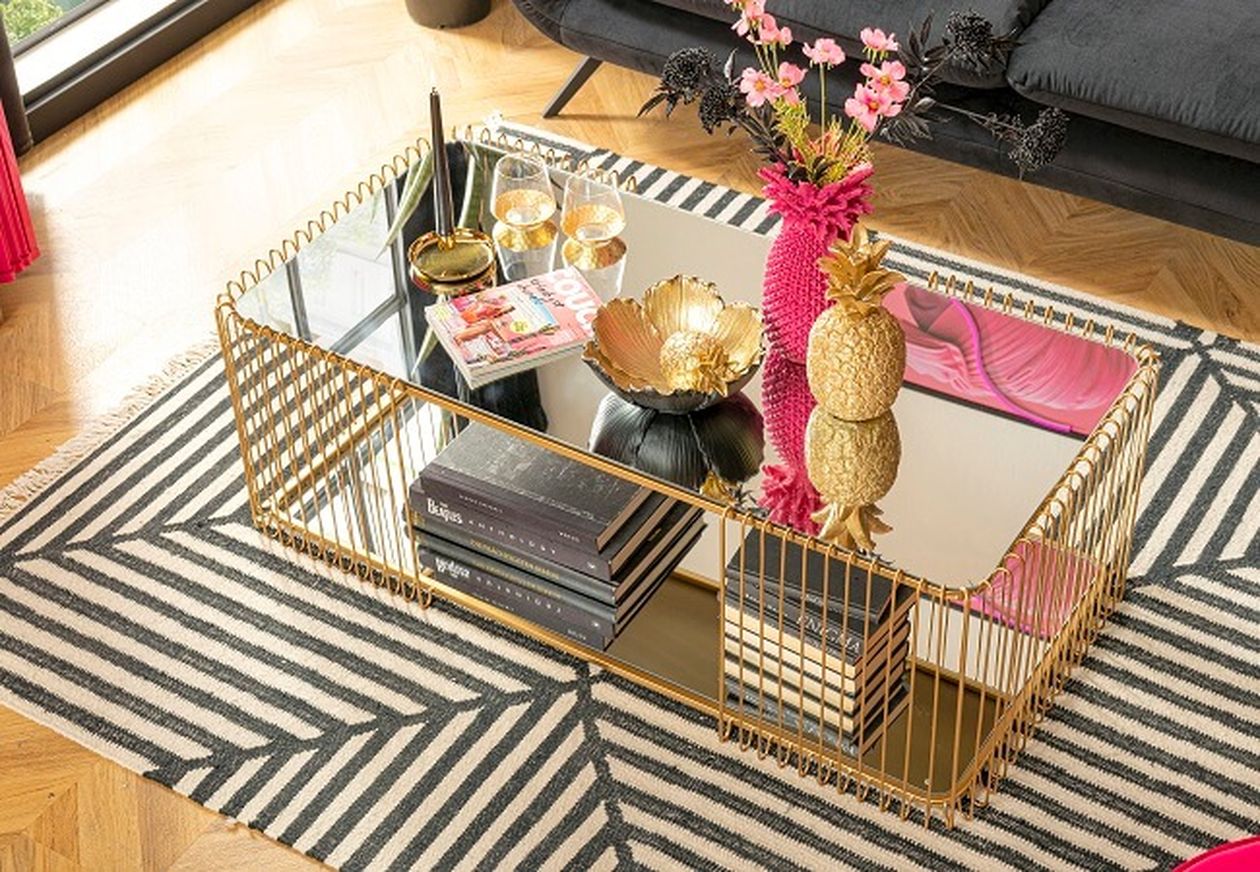 Ein schickes Wohnzimmer mit schwarzem Sofa, einer rosa Vase mit Blumen, goldenen Ananas und dekorativen Schalen auf einem Couchtisch aus reflektierendem Glas.