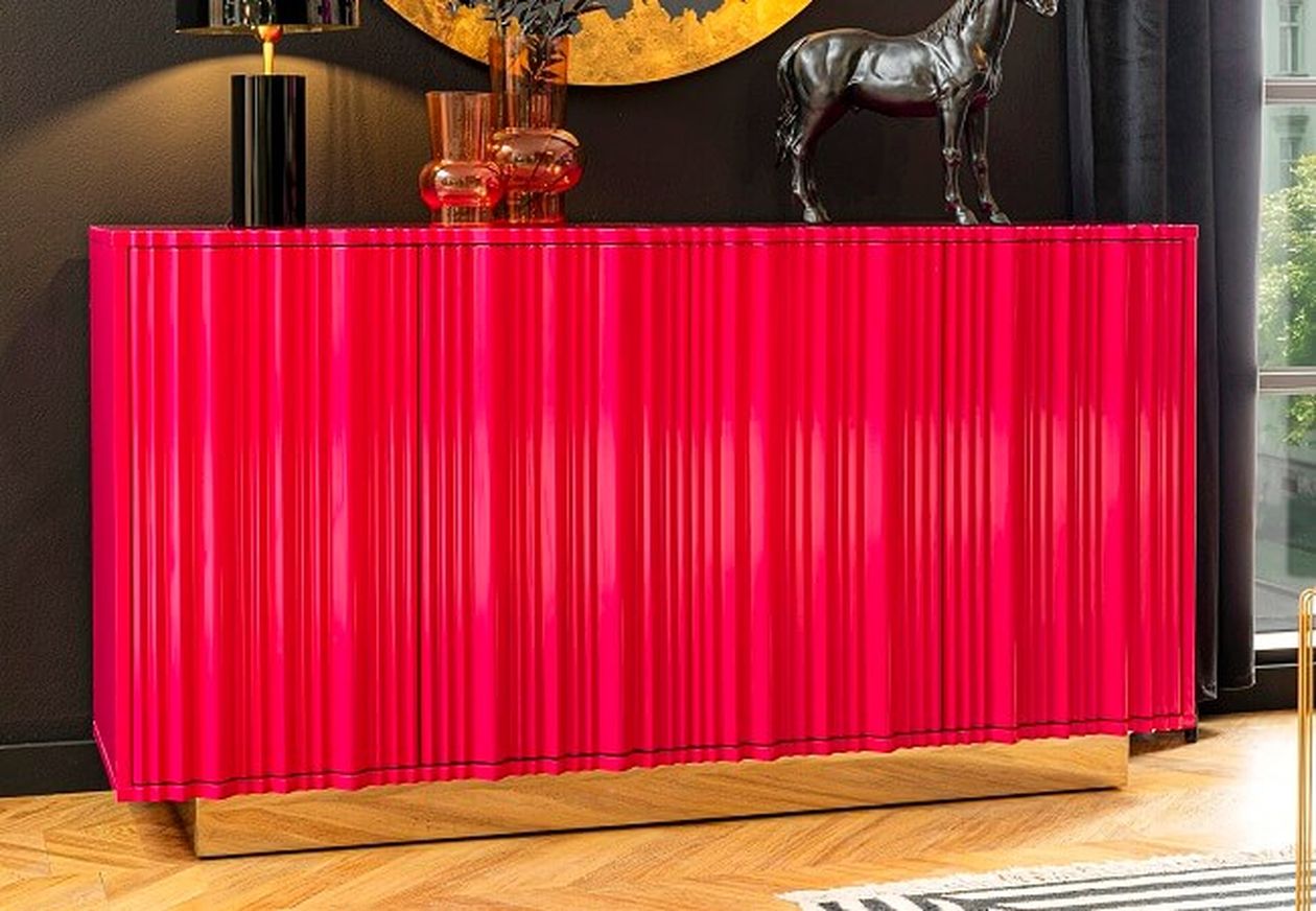 Ein leuchtend pinkfarbenes, modernes Sideboard mit gerippter Oberfläche, goldenem Sockel und dekorativen Elementen darauf, vor einer dunklen Wand.