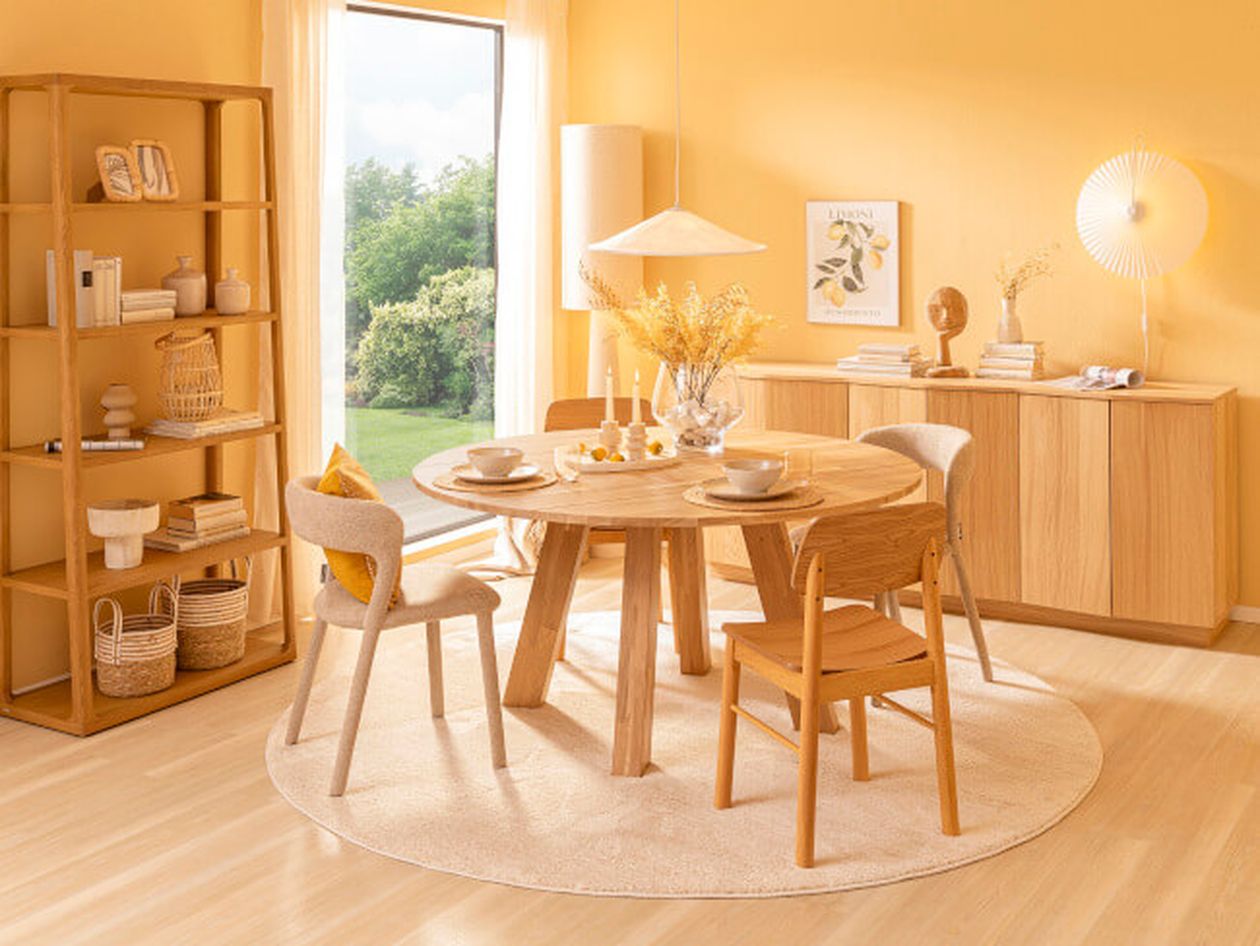 Heller Essbereich mit rundem Holztisch, vier Stühlen, Regal und Kommode. Gelbe Wandfarbe, großes Fenster, Dekoration mit Blumen und Kerzen.