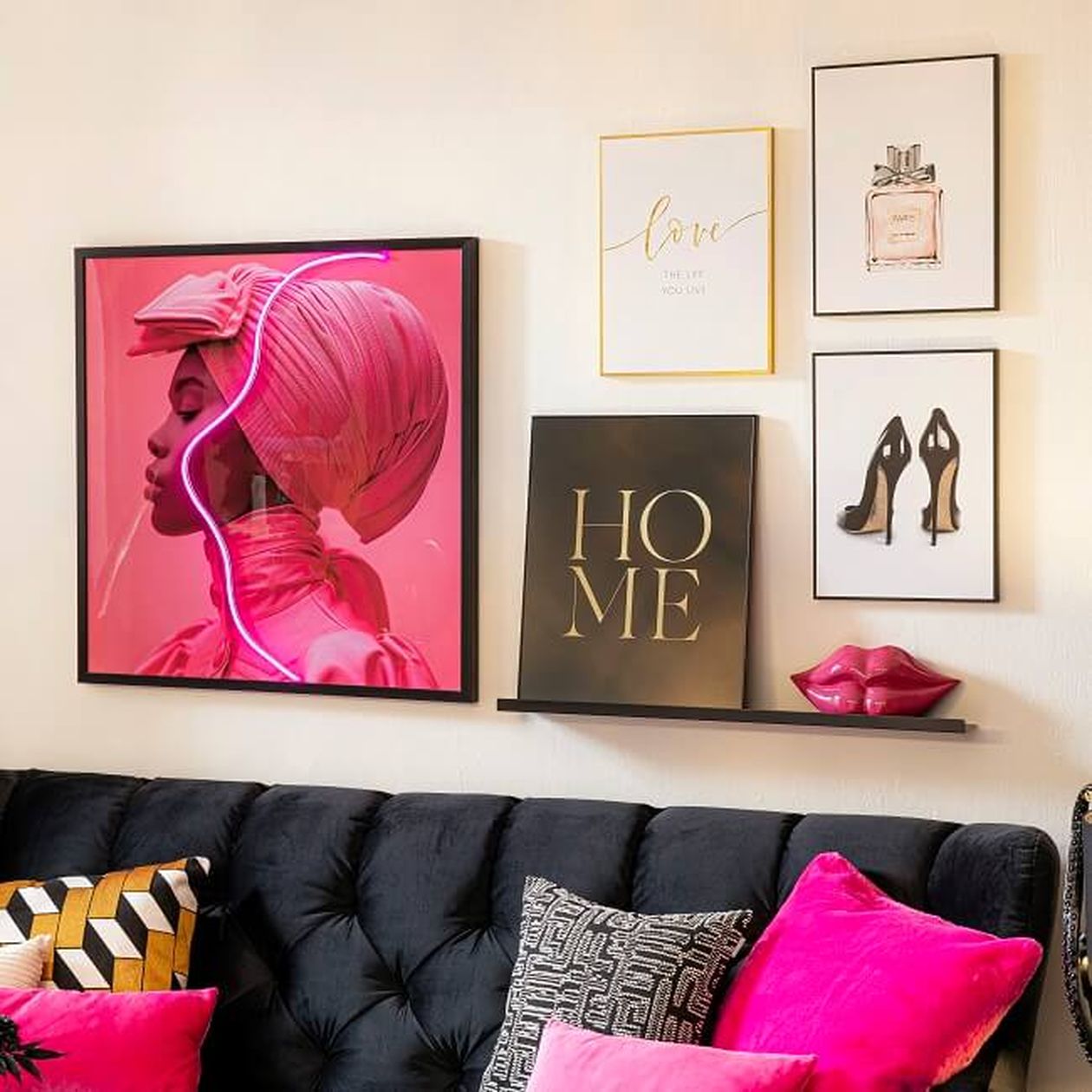 Wandbilder mit einem „HOME“-Schild, Illustrationen von Parfüm und Schuhen, rosa Lippen als Dekoration auf einem Regal, über einem schwarzen Sofa mit farbenfrohen Kissen.