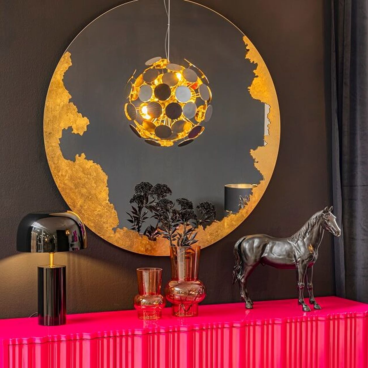 Ein leuchtend pinkfarbenes Sideboard mit einer modernen schwarzen Lampe, bernsteinfarbenen Vasen und einem runden goldenen Spiegel, der eine gold-schwarze Pendelleuchte an einer dunklen Wand reflektiert.