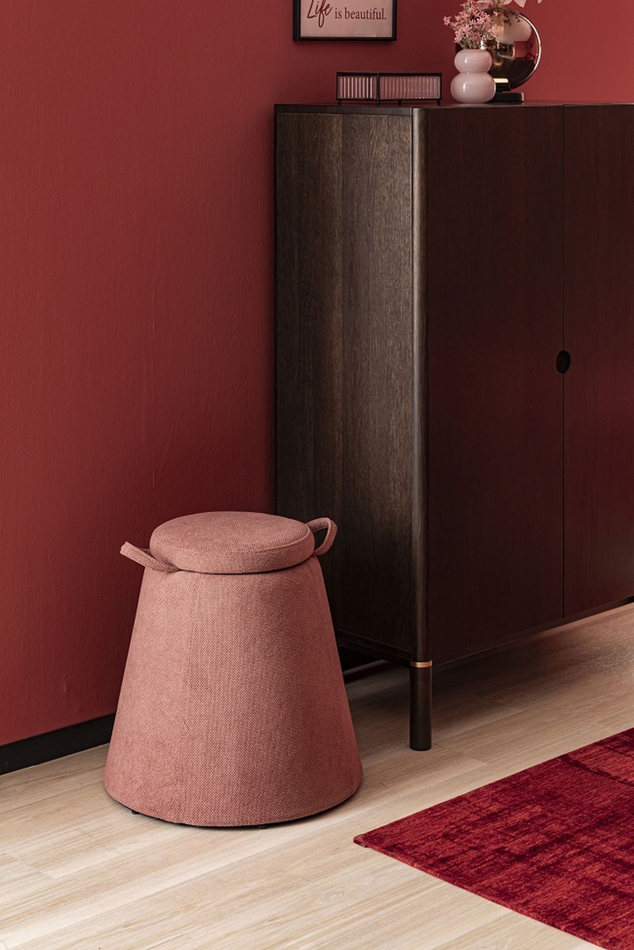 Roter Raum mit einem dunkelbraunen Schrank, einem rosanen Hocker und Dekoration. Ein Bild mit dem Schriftzug "Life is beautiful" hängt an der Wand.