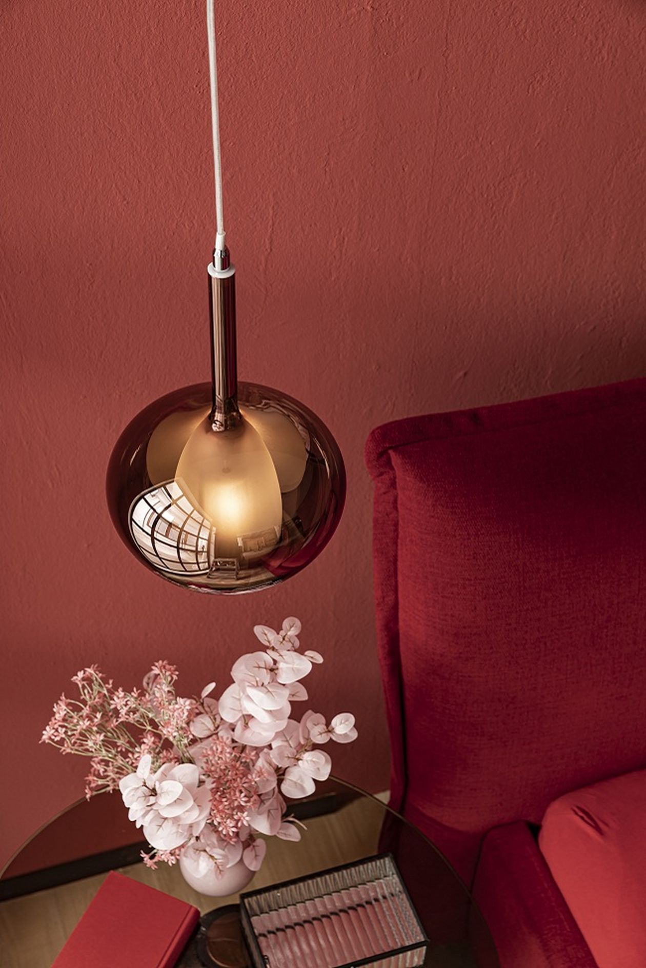 Kupferfarbene Hängeleuchte, rotes Sofa, runde Tischplatte mit rosa Blumen und Notizbuch, vor rotem Hintergrund.