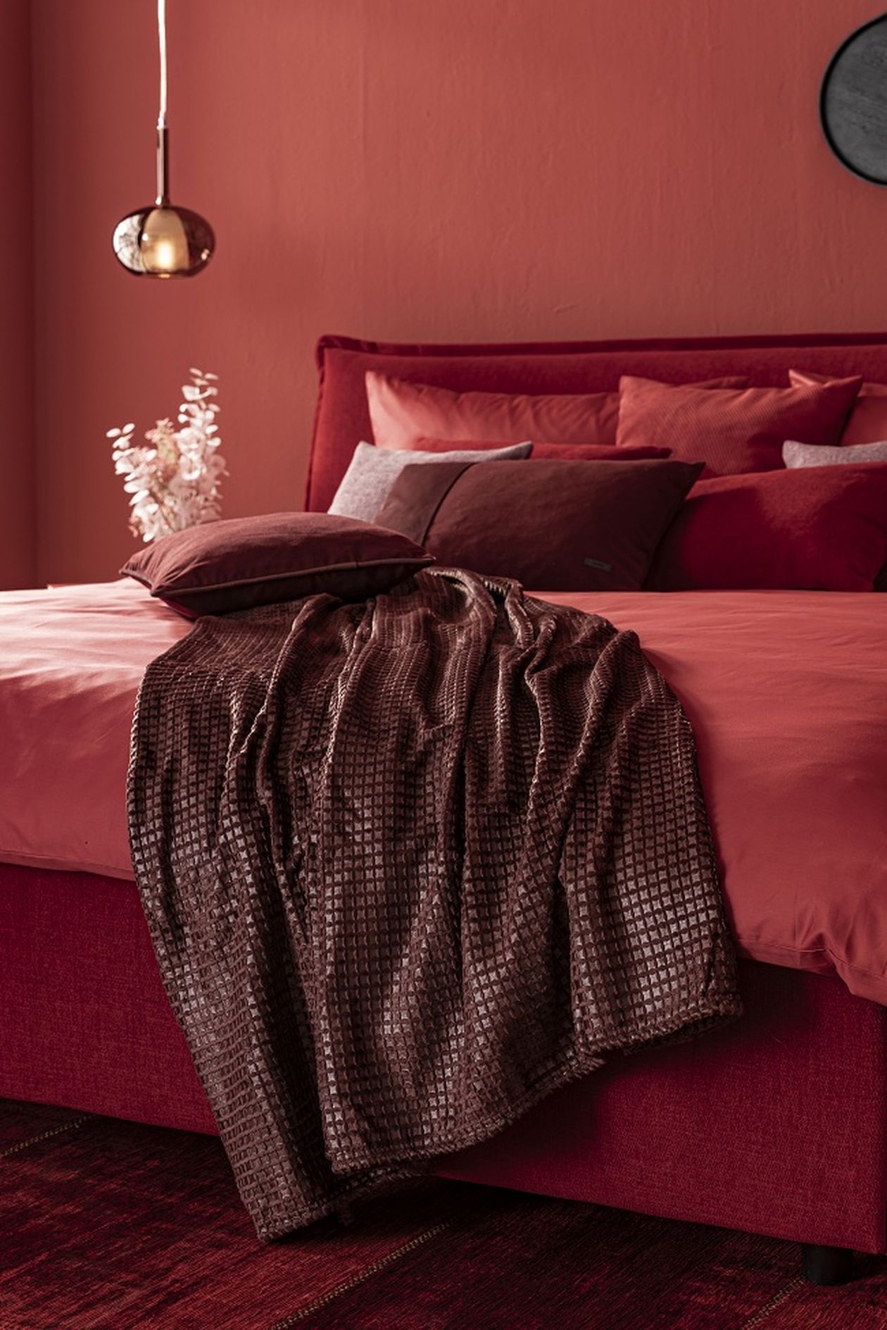 Elegantes Schlafzimmer in Rottönen mit rotem Bett, Kissen und Überwurf. Eine Hängelampe und Trockenblumen ergänzen das Ambiente.