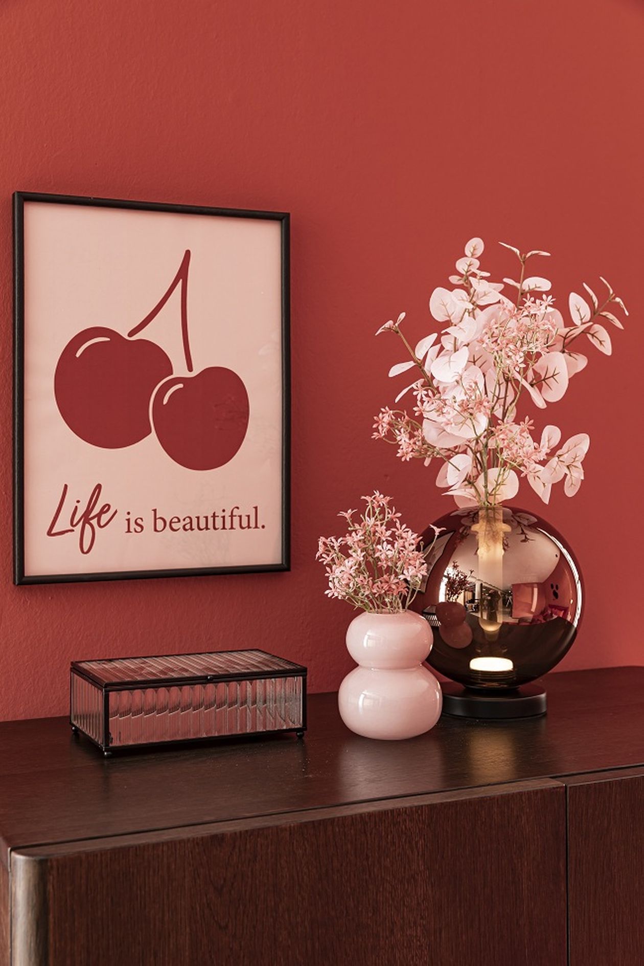 Roter Raum mit Bild von Kirschen und Text "Life is beautiful", Vase mit rosa Blumen auf einem dunklen Holztisch.