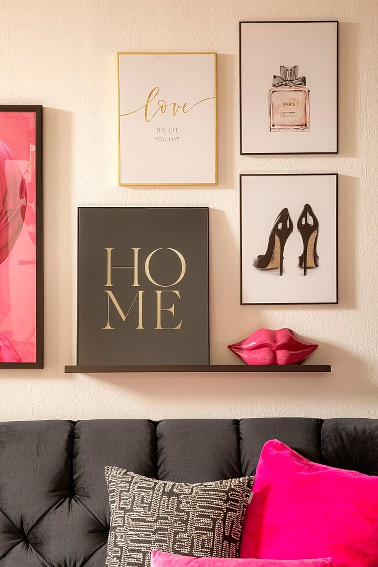 Wandbilder mit einem „HOME“-Schild, Illustrationen von Parfüm und Schuhen, rosa Lippen als Dekoration auf einem Regal, über einem schwarzen Sofa mit farbenfrohen Kissen.