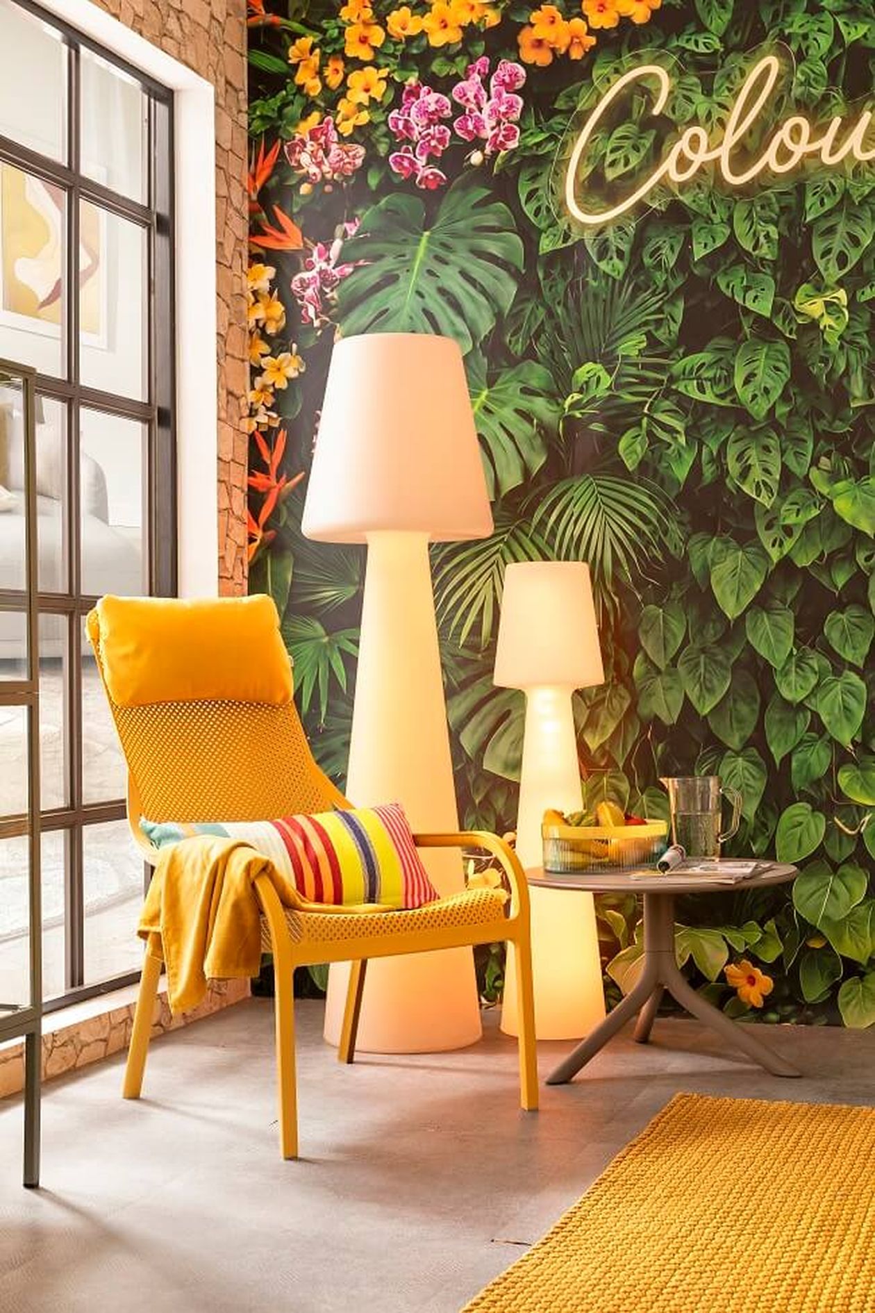 Gemütlicher Outdoor-Bereich mit gelbem Sessel, gestreiftem Kissen, Stehlampen und grüner Blättertapete. Regale mit Pflanzen und Dekorationen stehen an einem großen Fenster.