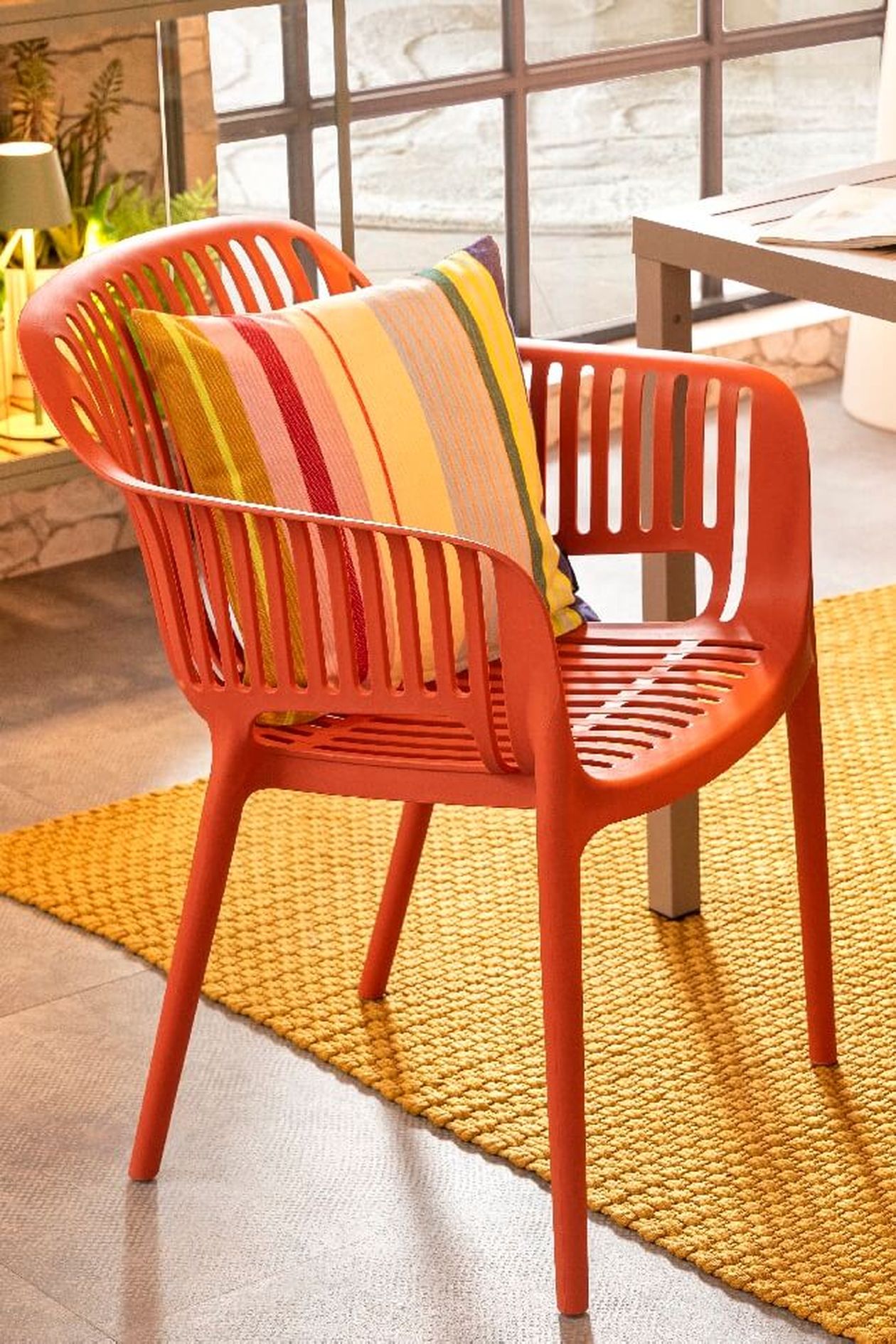 Ein rot-orangefarbener Plastikstuhl mit gestreiftem Kissen auf einem gelben Webteppich, neben einem Tisch an großen Fenstern.
