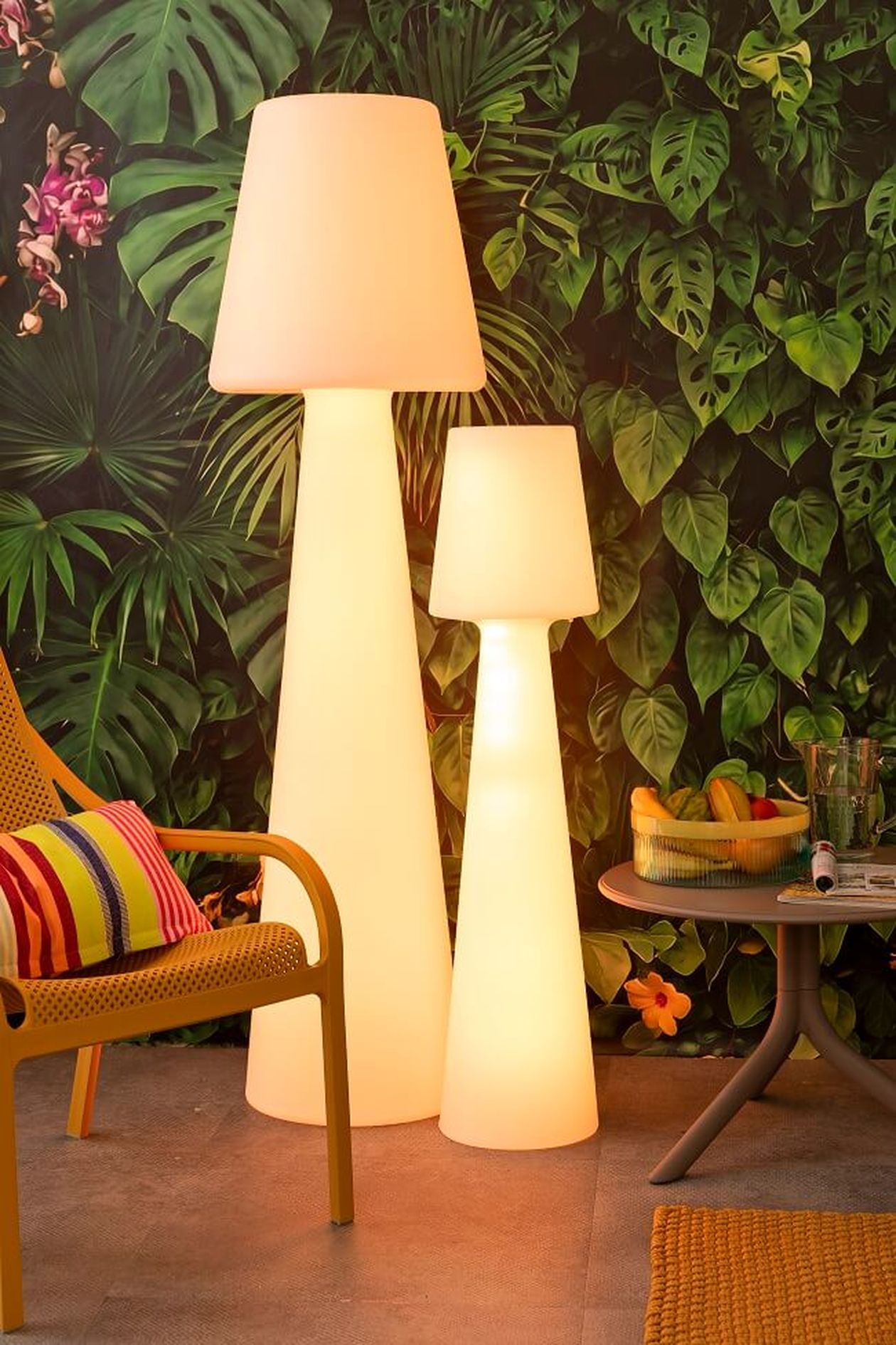 Zwei beleuchtete Stehlampen vor einer blattreichen, tropischen Tapete, daneben ein Korbsessel und ein kleiner Tisch mit Getränken und Obst.