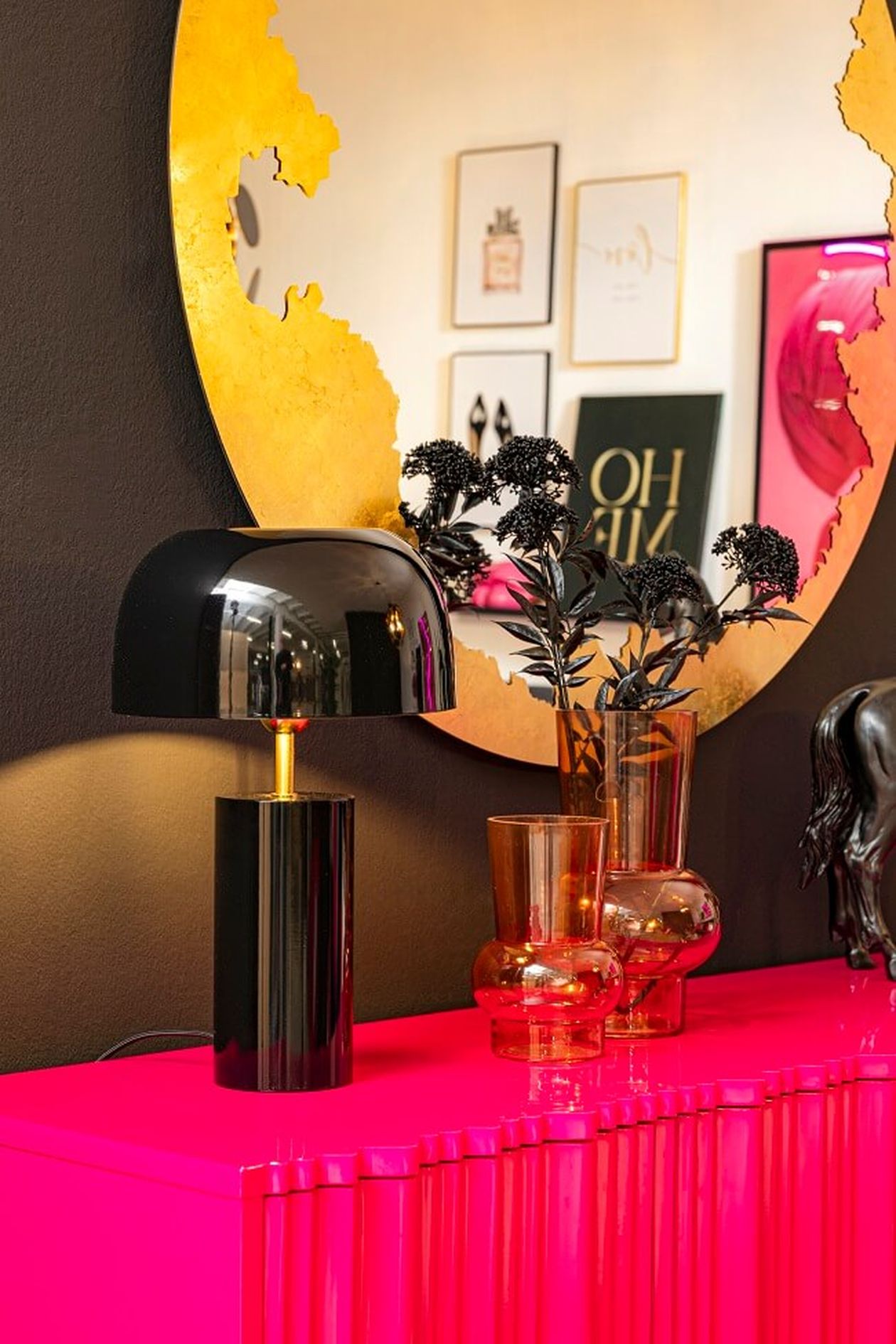 Ein leuchtend pinkfarbenes Sideboard mit einer modernen schwarzen Lampe, bernsteinfarbenen Vasen und einem runden goldenen Spiegel, der gerahmte Kunstwerke an einer dunklen Wand reflektiert.