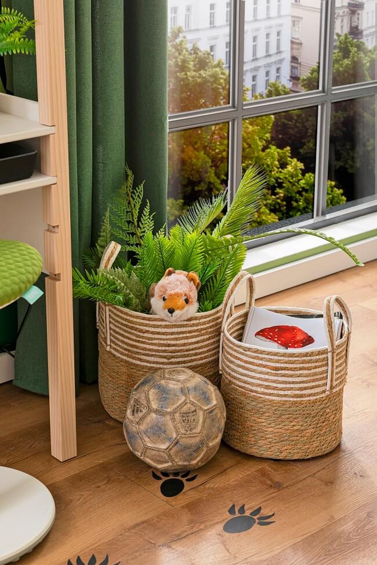 Zwei geflochtene Körbe mit einem Plüschtier, Pflanzen und Büchern stehen neben einem Fenster. Auf einem Holzboden mit Pfotenabdruck-Aufklebern liegt ein Fußball.