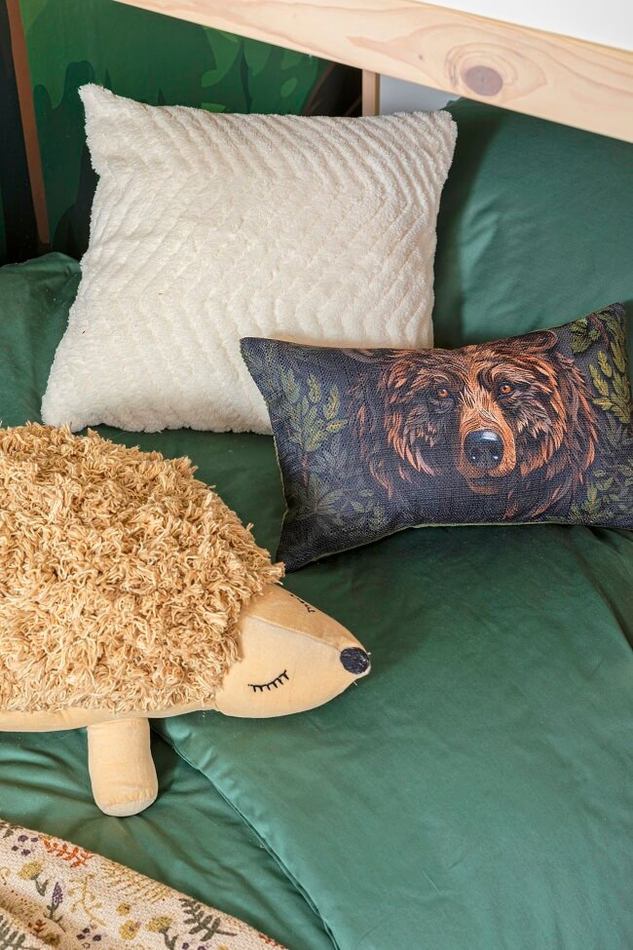 Bett mit grünen Laken, einem weißen, strukturierten Kissen, einem igelförmigen Plüschkissen und einem Kissen mit einer Bärenabbildung.