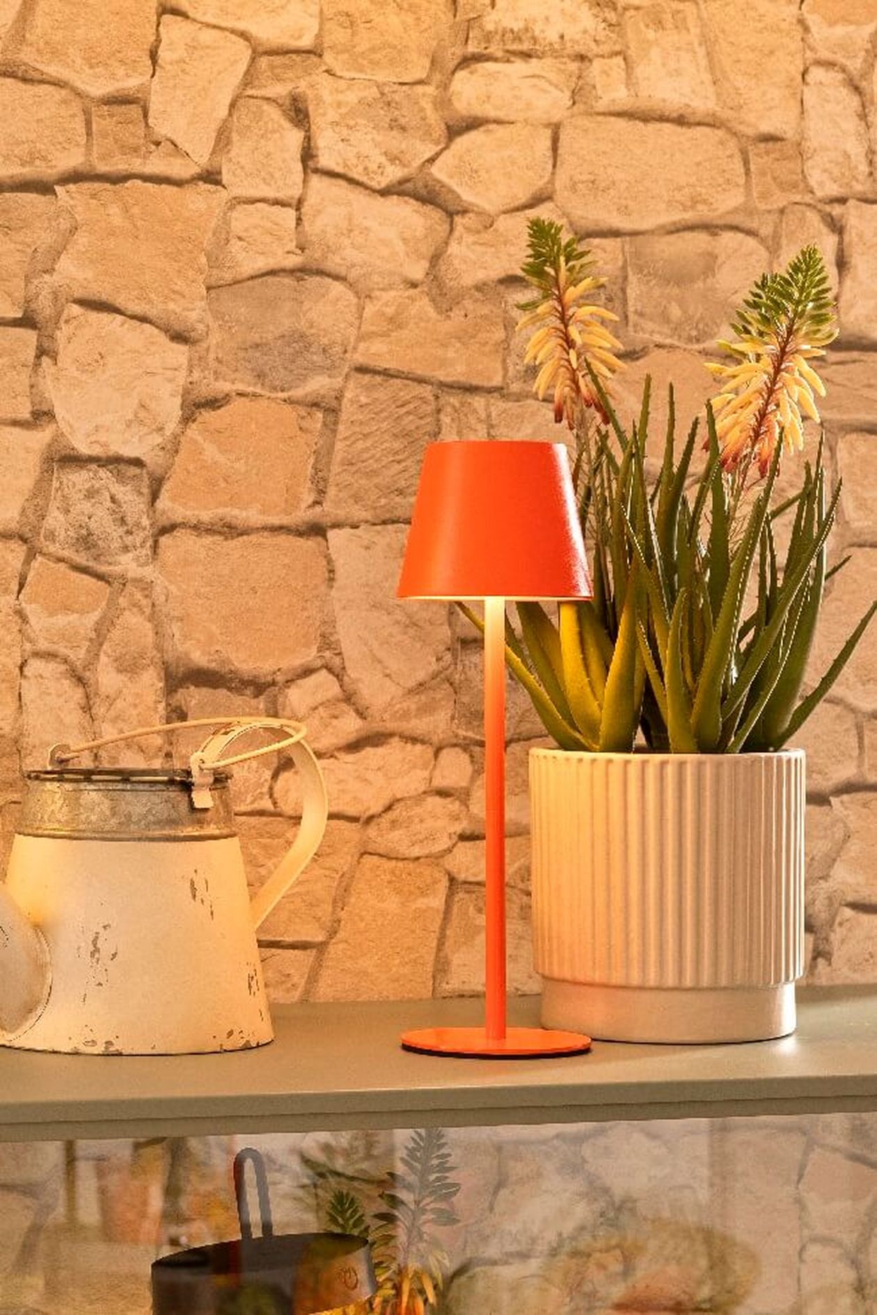 Eine orangefarbene Tischlampe, eine weiße Gießkanne und eine Topfpflanze mit hohen Blüten auf einem Regal vor einer Wand mit Steinstruktur.