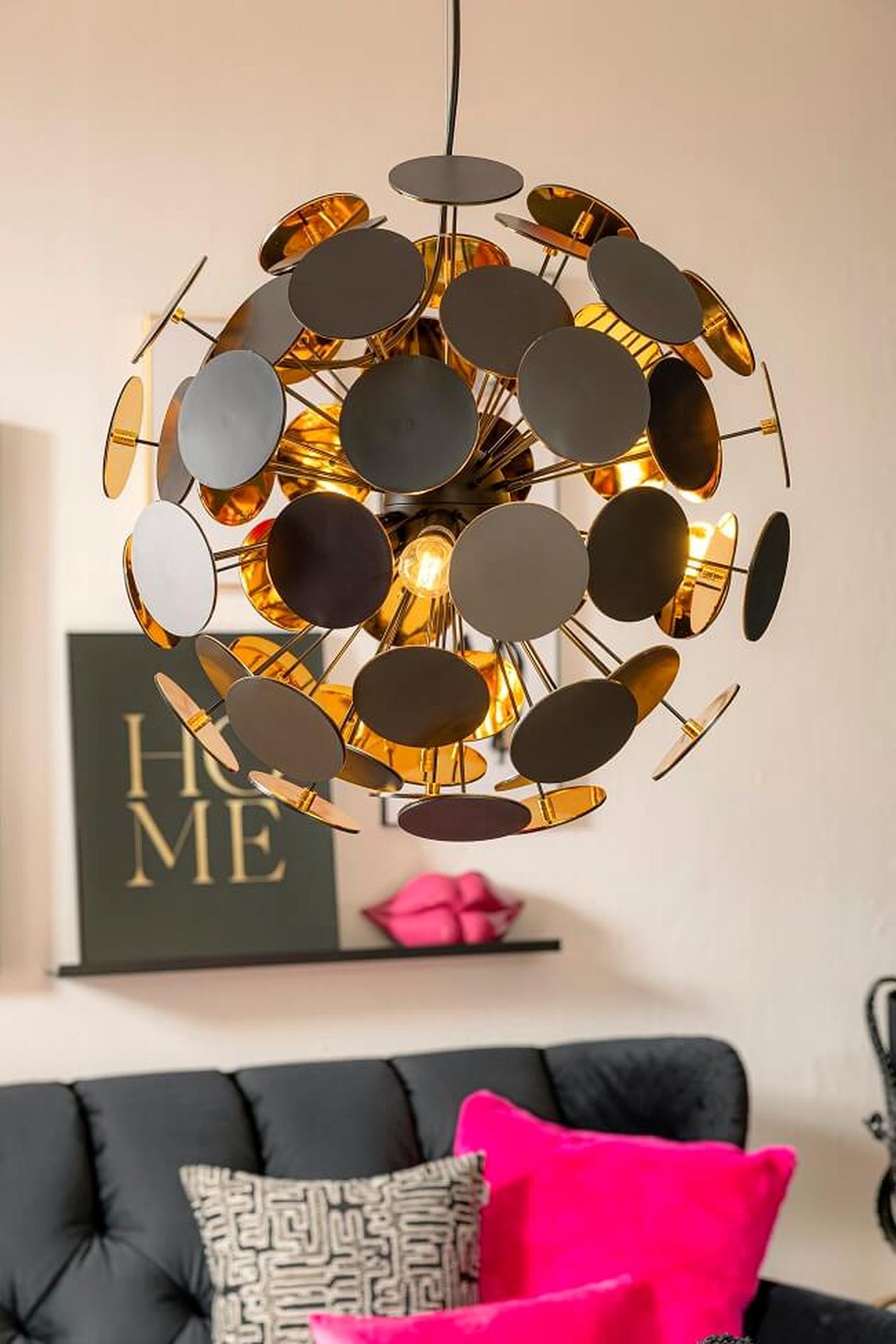 Ein moderner, kugelförmiger Kronleuchter mit goldenen Akzenten und schwarzen Scheiben hängt über einem schwarzen Sofa mit rosa und gemusterten Kissen. In der Nähe befindet sich ein Wandbild.