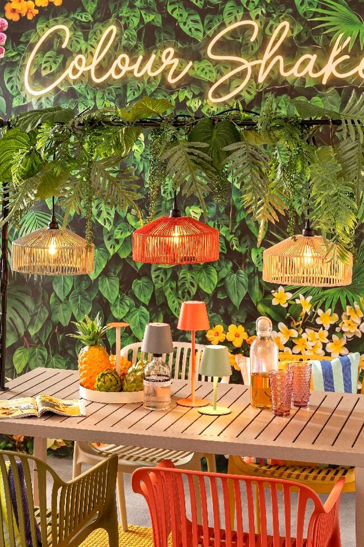 Lebhafte Terrasse im tropischen Stil mit farbenfrohen Pendelleuchten, üppigem Grün, Rattanstühlen und einem mit Obst und Dekorationsgegenständen gedeckten Tisch.