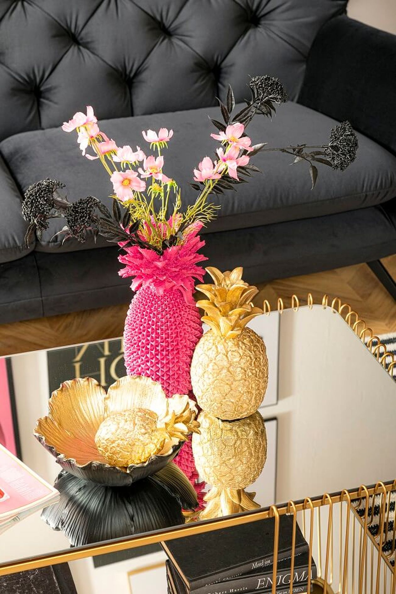Ein schickes Wohnzimmer mit schwarzem Sofa, einer rosa Vase mit Blumen, goldenen Ananas und dekorativen Schalen auf einem Couchtisch aus reflektierendem Glas.
