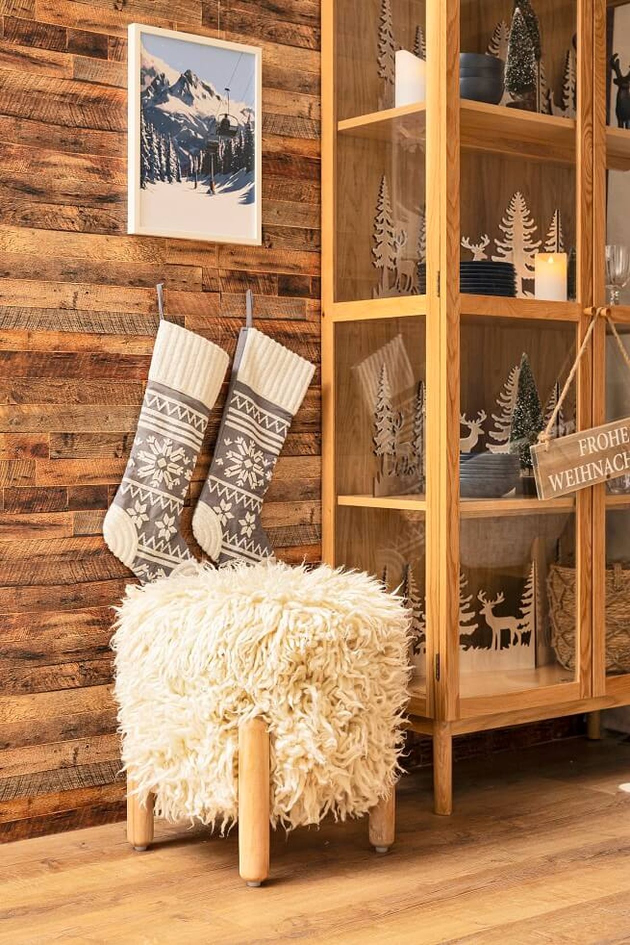 Gemütliches Zimmer mit Holzwänden, festlichen Strümpfen, einem flauschigen Hocker und einem Schrank mit Weihnachtsdeko. Darüber hängt ein Foto eines verschneiten Berges.