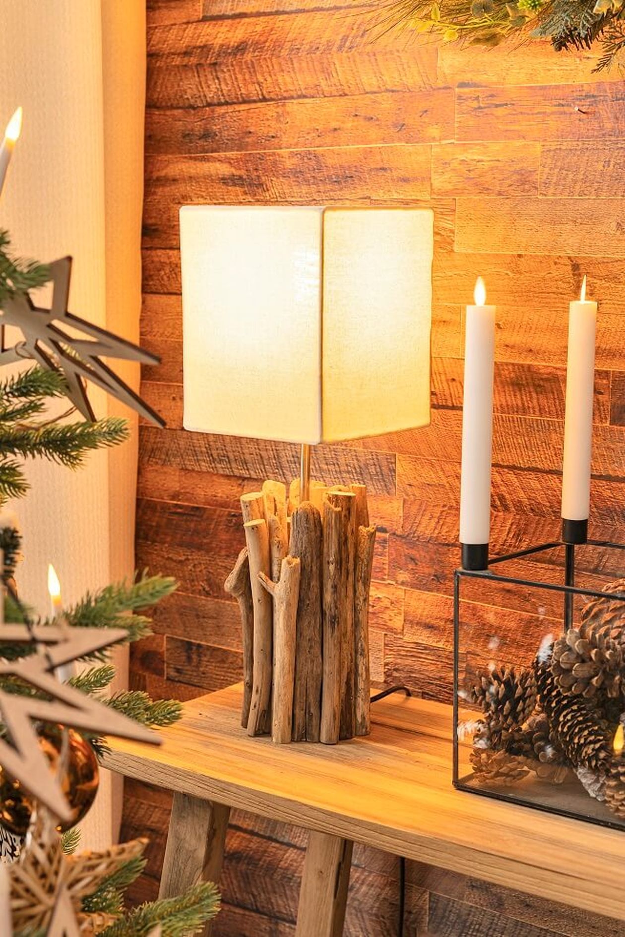 Eine rustikale Lampe mit einem Sockel aus Treibholz und einem weißen Schirm steht auf einem Holztisch neben brennenden Kerzen und Tannenzapfen vor einer Holzwand.