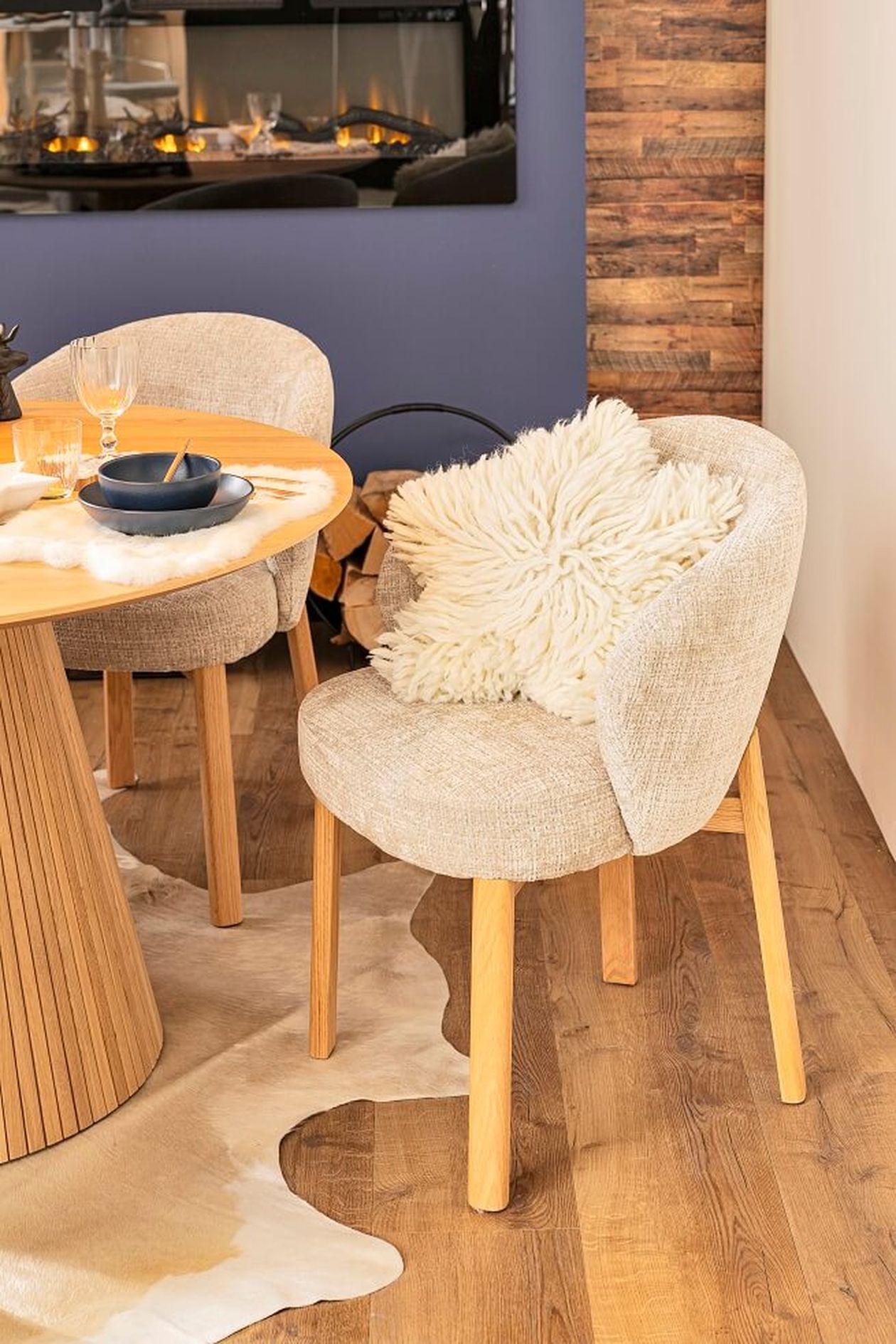 Gemütlicher Essbereich mit einem runden Holztisch, gepolsterten Stühlen, flauschigen weißen Kissen und einem Kamin im Hintergrund.