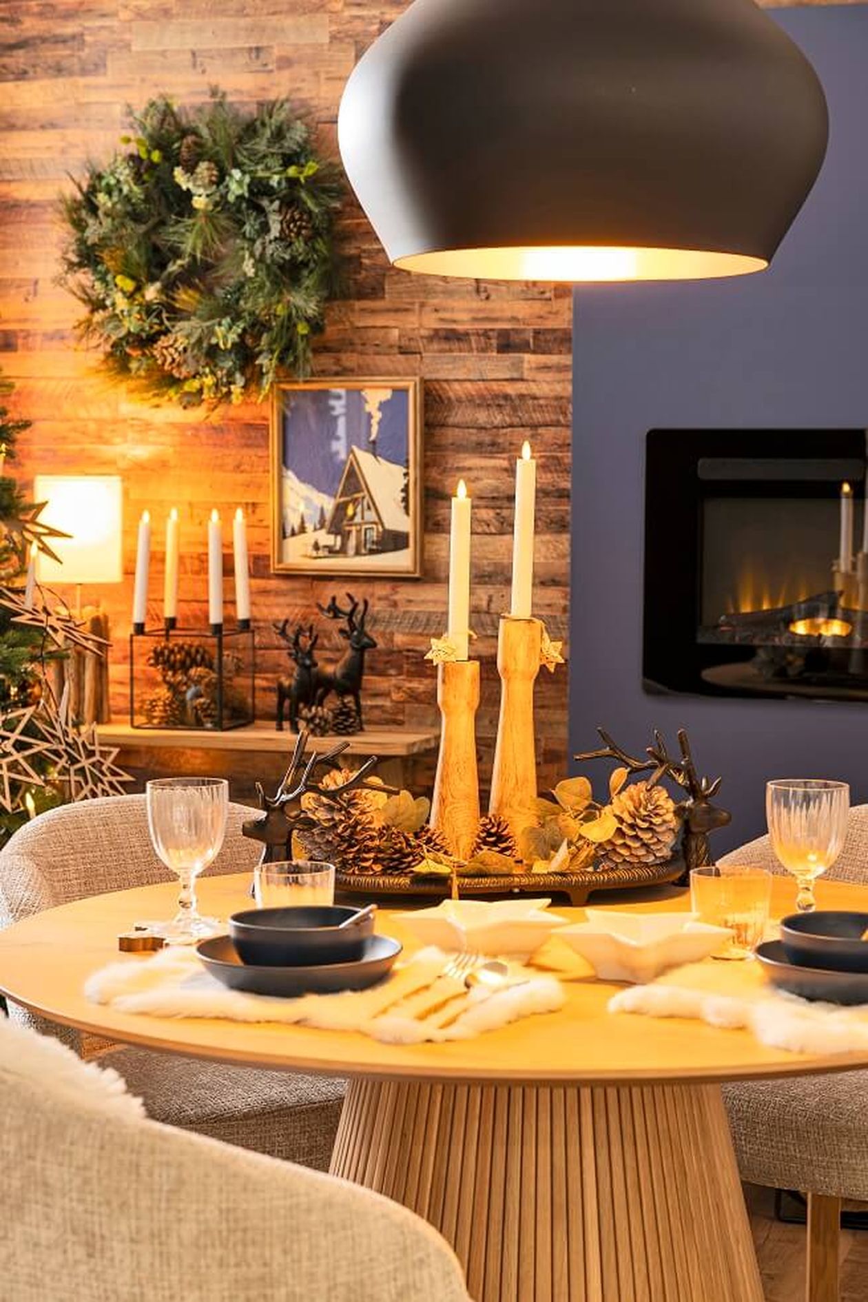 Gemütlicher Essbereich mit rundem Tisch, Kerzen, Tannenzapfen und einem Kranz an einer holzgetäfelten Wand. Ein Kamin und Weihnachtsdeko sorgen für Wärme.