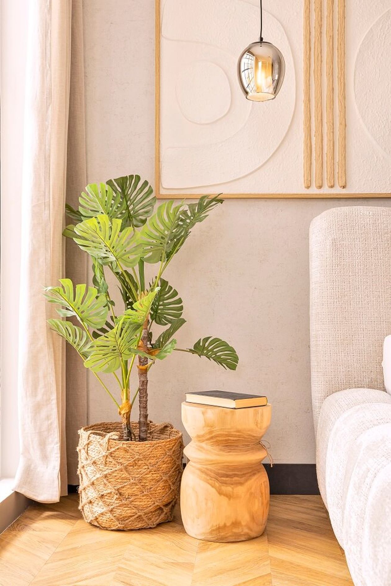 Künstliche, grüne Monstera-Pflanze in einem Blumentopf aus Korbgeflecht neben einem runden Hocker aus Holz.