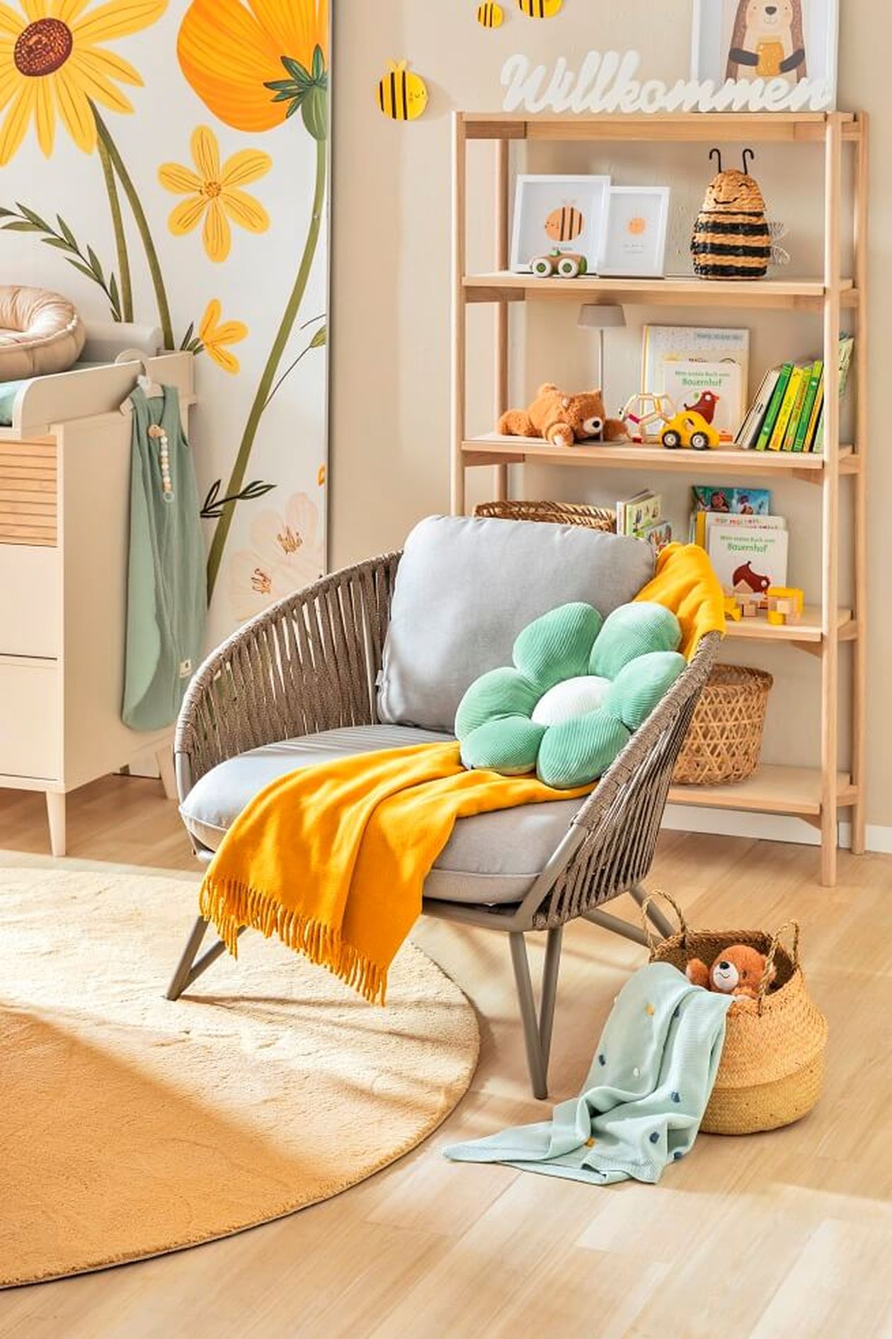 Gemütlicher Stillsessel mit gelber Kucheldecke, grünem Blumenkissen und einem Bücherregal im Hintergrund.