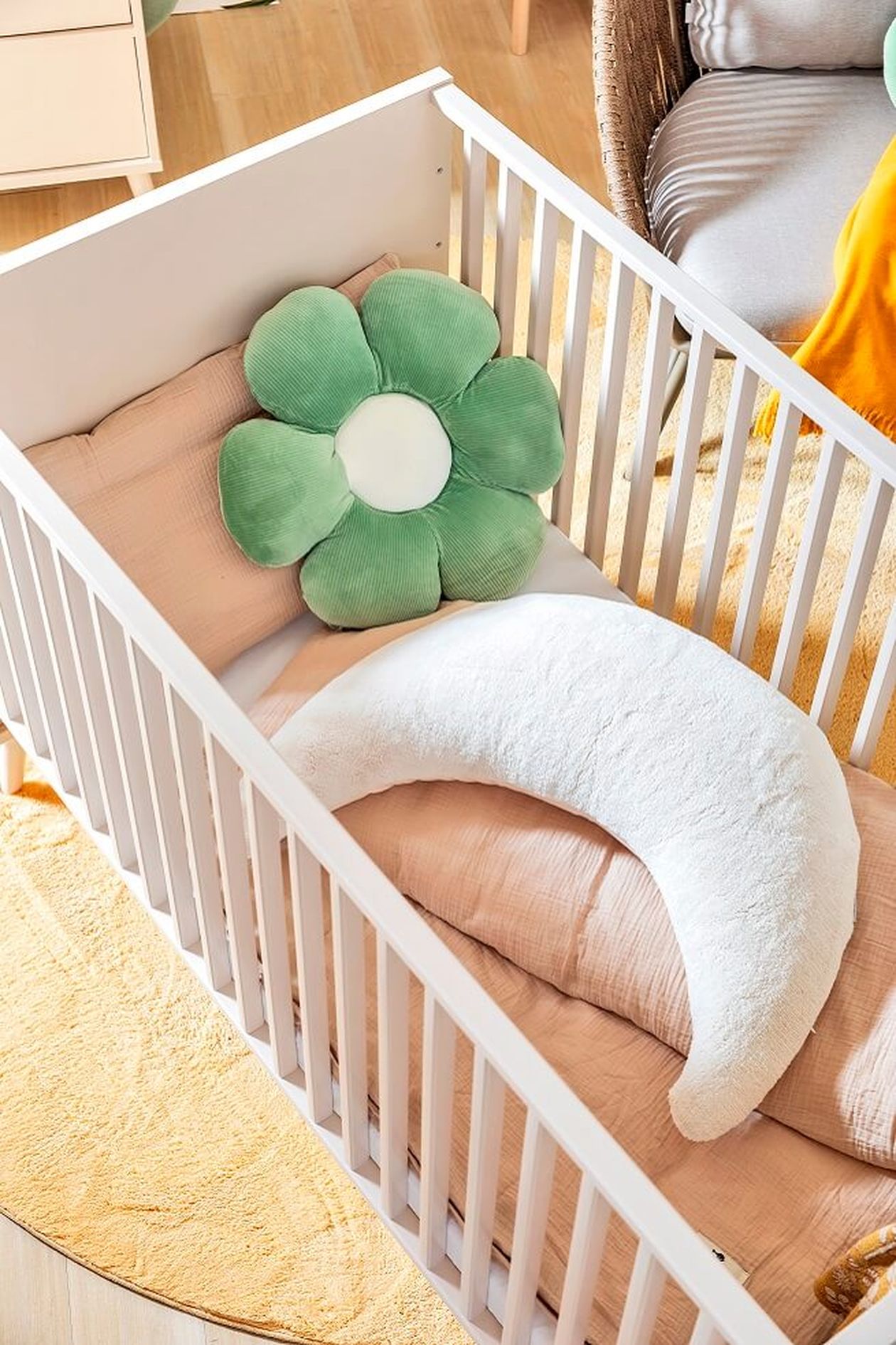 Baby-Gitterbett von oben fotografiert mit beiger Bettwäsche und grünem Kissen in Blumen-Form.