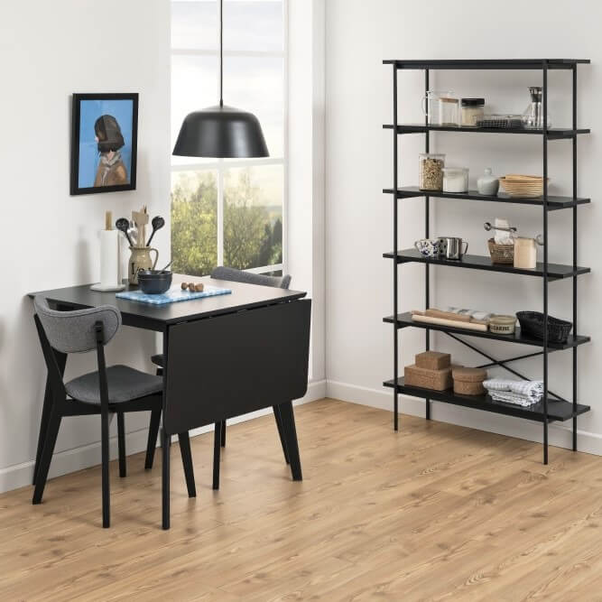 Schwarzer Klapptisch in eingeklapptem Zustand steht in einem Esszimmer. Dahinter steht Wandregal in gleichem Design.