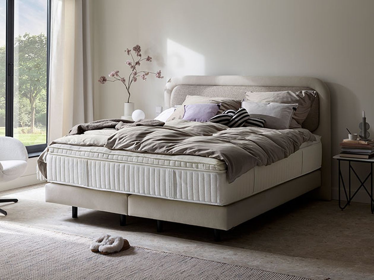 Ein gemütliches Schlafzimmer mit ordentlich gemachtem Bett, sanfter Beleuchtung und Kissen, in der Nähe eines großen Fensters mit Blick ins Grüne.