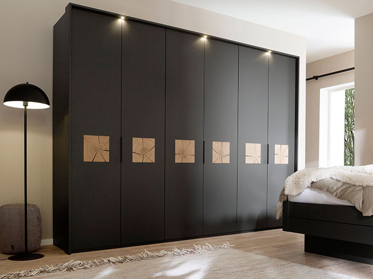 Ein modernes Schlafzimmer besticht durch einen eleganten schwarzen Kleiderschrank mit Holzelementen, ein gemütliches Bett, einen grauen Hocker und eine schwarze Stehlampe – alles auf Holzboden.