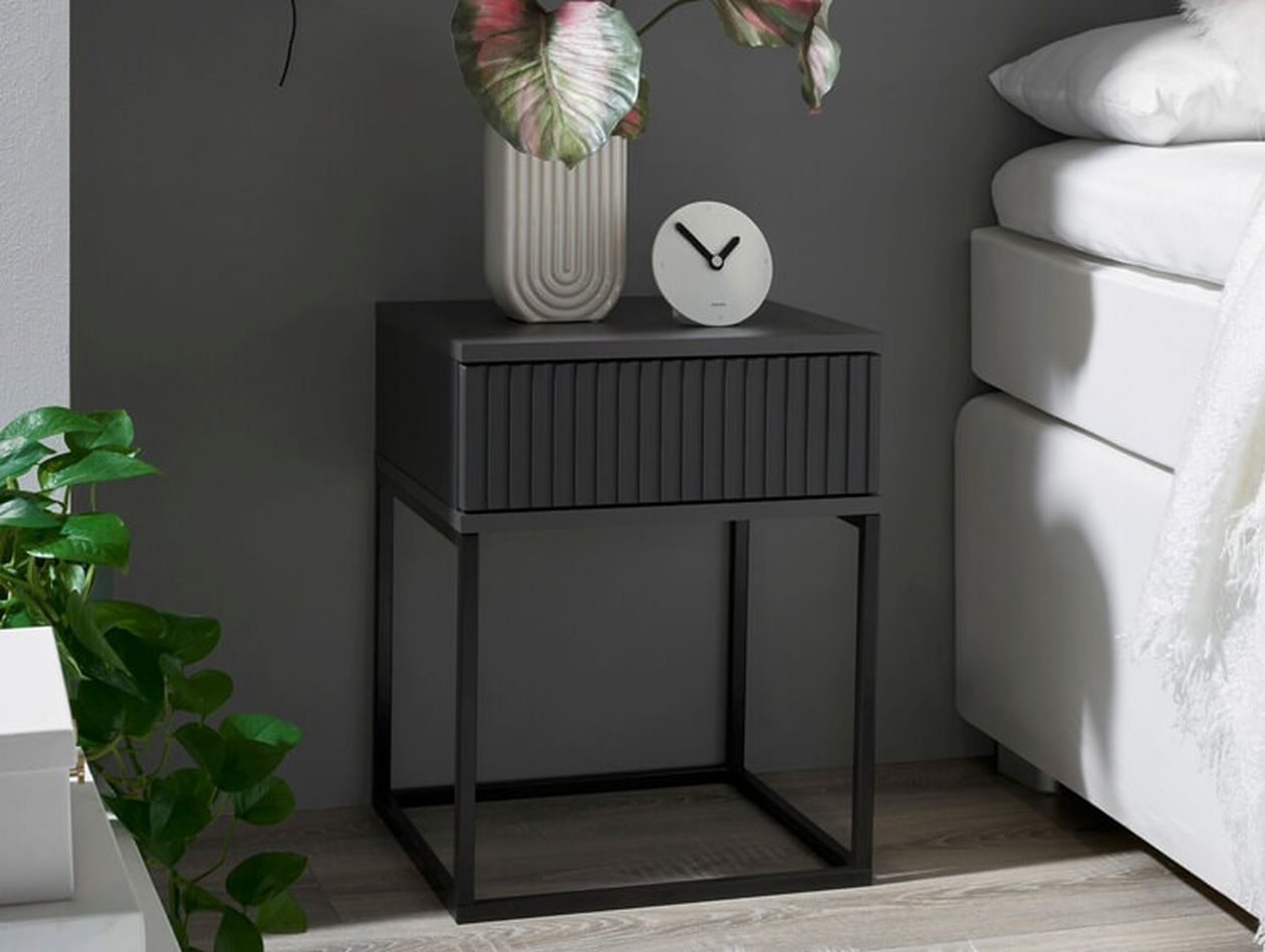 Schwarzer Nachttisch mit geriffelter Schubladenfront und schlankem Metallgestell neben einem weißen Bett. Auf dem Nachttisch stehen eine dekorative Vase mit Pflanze und eine runde weiße Uhr. Im Hintergrund eine dunkelgraue Wand und eine grüne Zimmerpflanze.