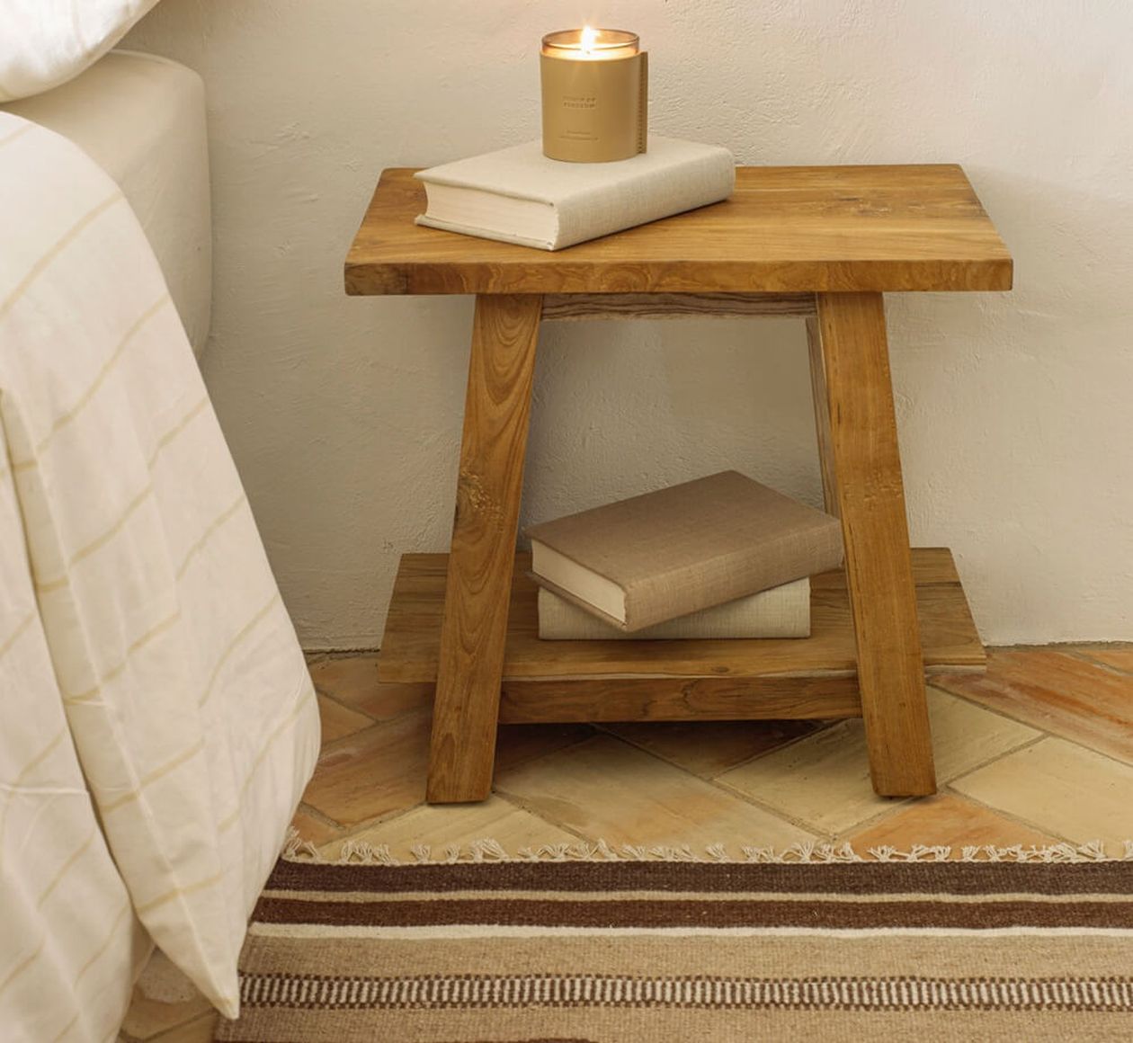 Nachttisch aus Holz mit einer brennenden Kerze und zwei Büchern darauf und einem weiteren Buch auf dem unteren Regal, neben einem Bett mit einer weißen Decke.