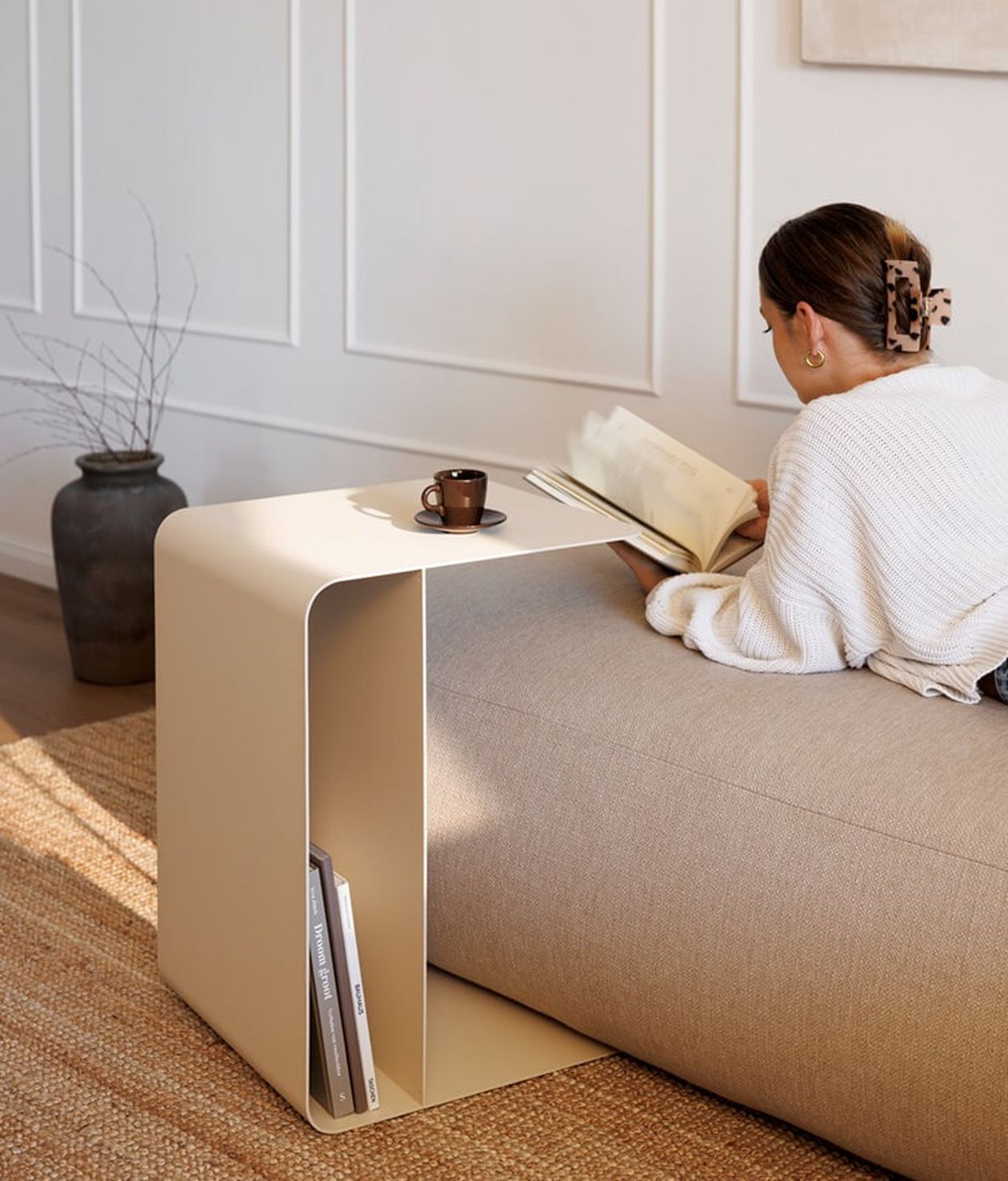Eine Frau liest auf einem beigen Sofa neben einem modernen Beistelltisch mit Kaffeetasse und Büchern in einem sanft beleuchteten Raum mit einer Vase und einem strukturierten Teppich.