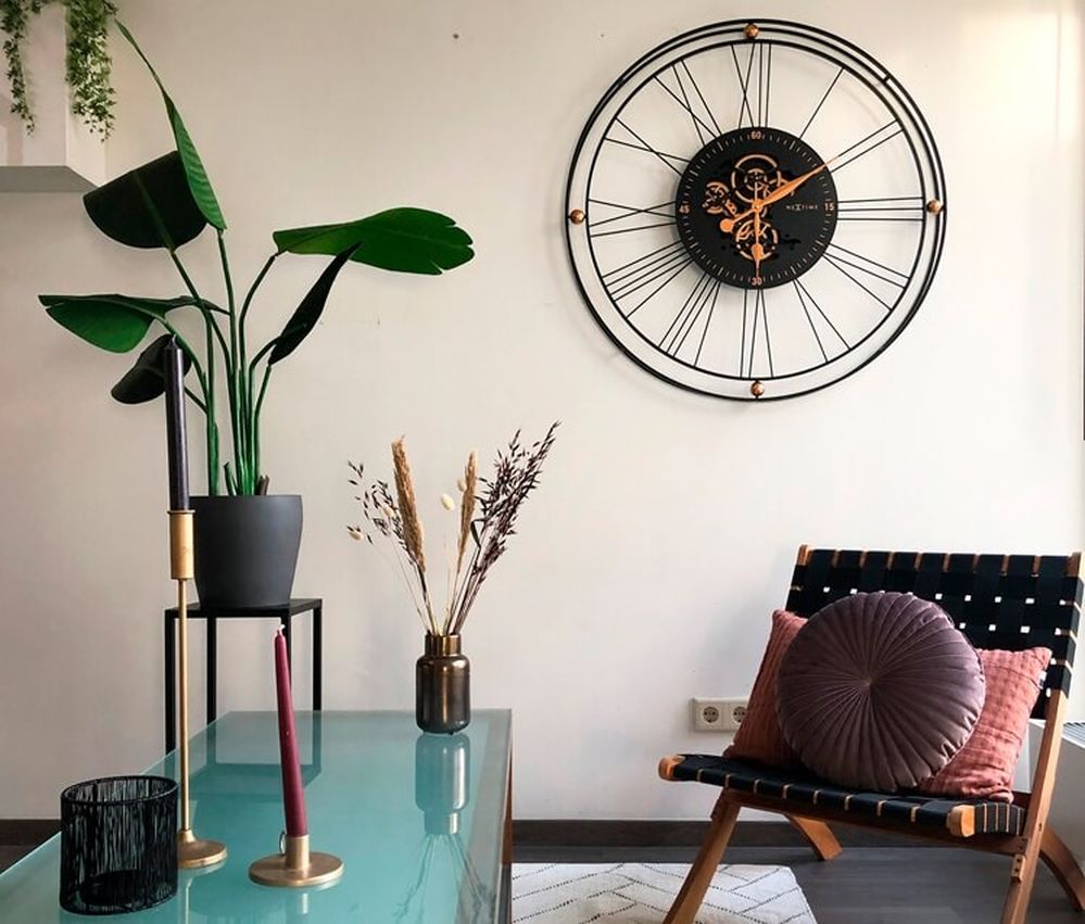 Modernes Wohnzimmer mit einer großen Wanduhr, einer Topfpflanze, einem Glastisch mit Kerzen und einem Stuhl mit Kissen vor einer weißen Wand.