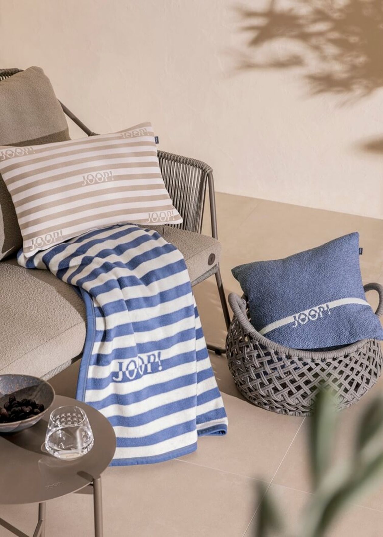 Blau-weiß gestreifte JOOP!-Decke auf einem Outdoor-Sessel mit Seilgeflecht. Daneben ein blauer JOOP!-Kissenbezug in einem Korb und beige-weiß gestreifte Kissen.