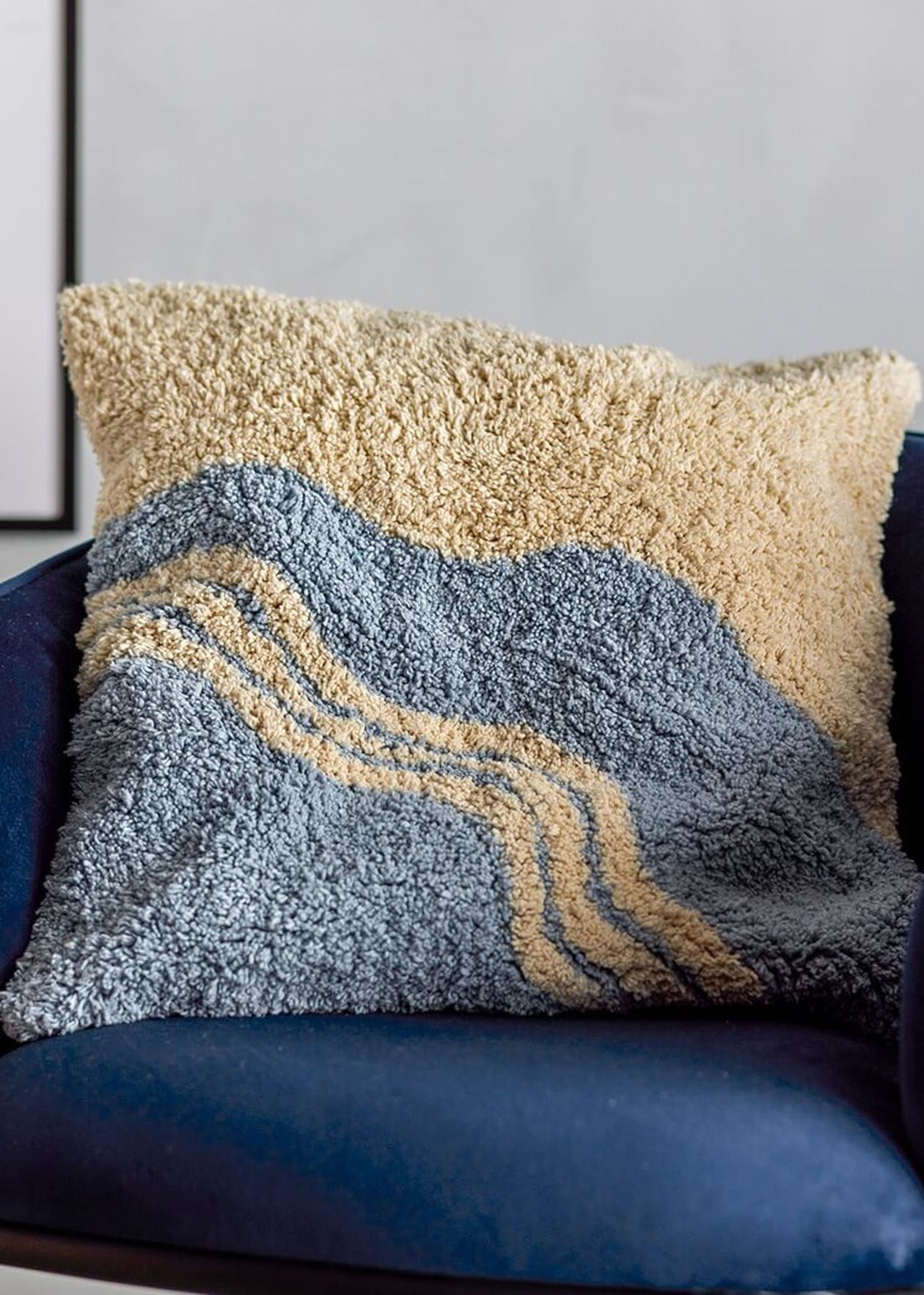 Flauschiges Dekokissen mit wellenförmigem Muster in Blau-Grau und Beige auf einem dunkelblauen Samtsessel.