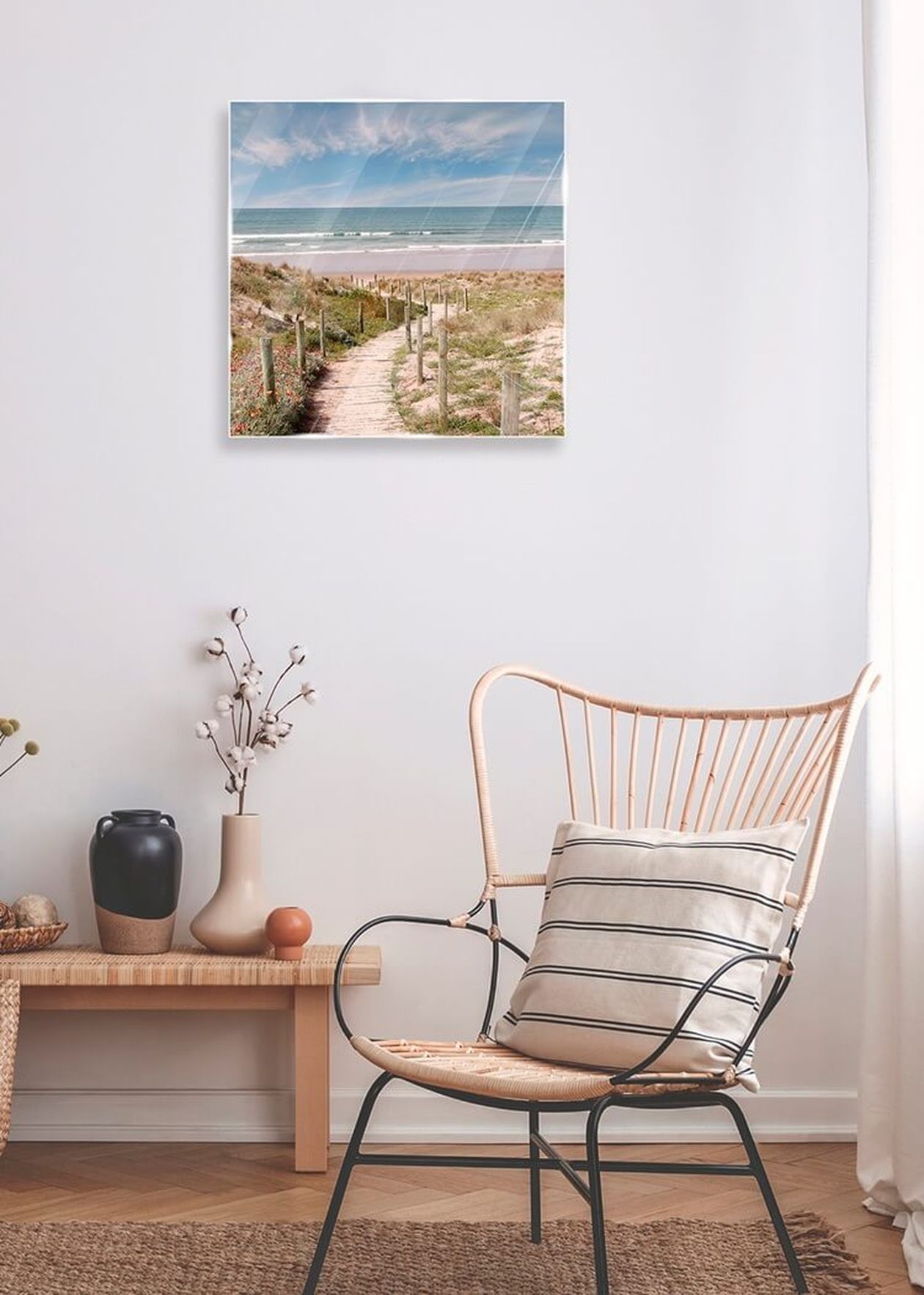 Quadratisches Glasbild mit einem Strandweg durch Dünen zum Meer an einer weißen Wand. Darunter ein Rattanstuhl mit gestreiftem Kissen und eine Bank mit Vasen und Deko.