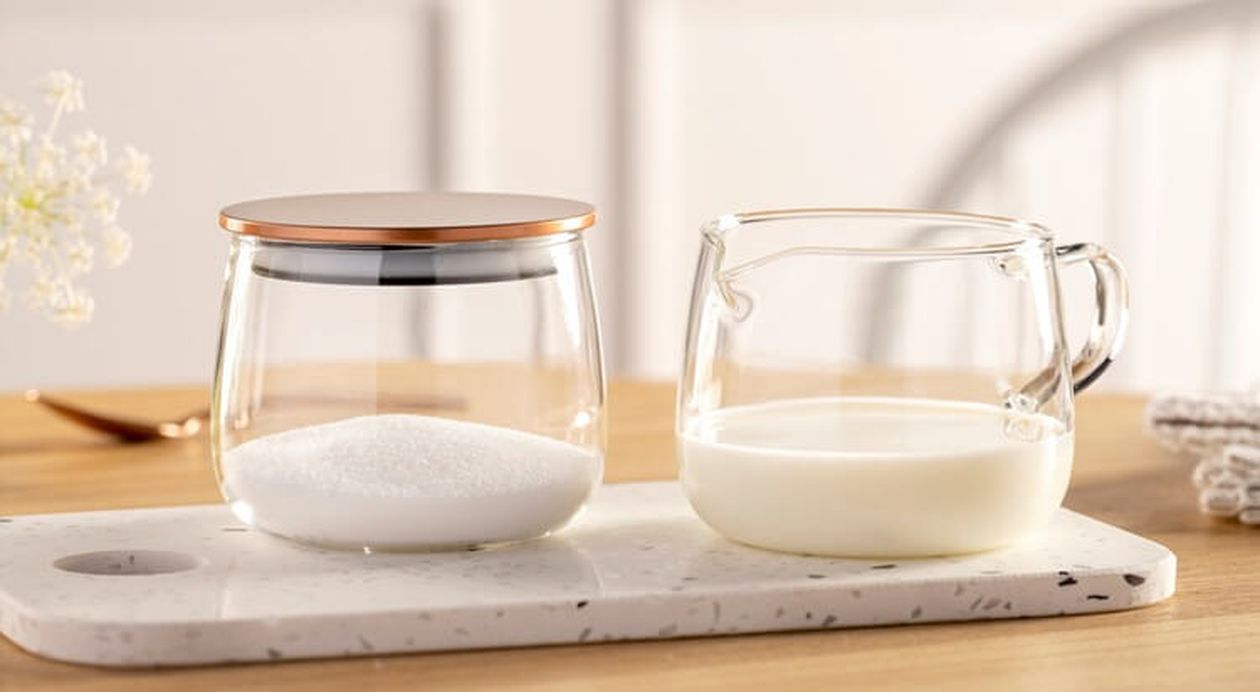 Gläser mit Zucker und Milch stehen auf einem Marmorschneidebrett auf einem Holztisch, im Hintergrund sind ein verschwommener weißer Stuhl und eine Wand zu sehen.