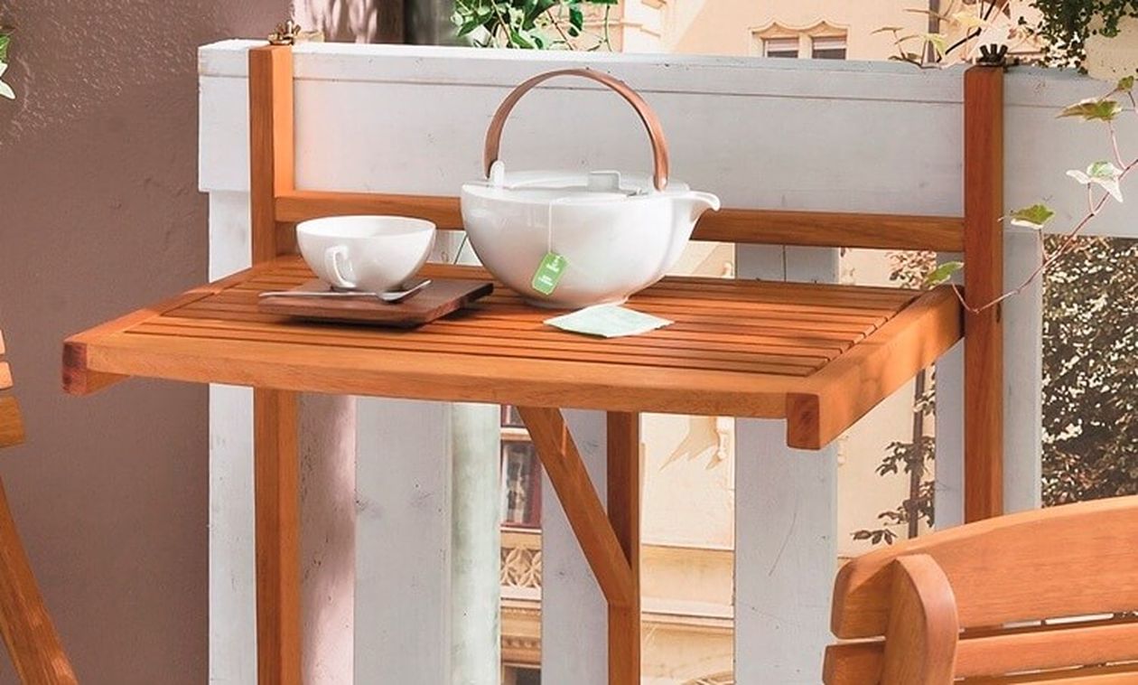 Klapptisch aus Holz auf einem Balkon mit einer weißen Teekanne, einer Teetasse und einer Untertasse. Weißes Geländer und Gebäude im Hintergrund.
