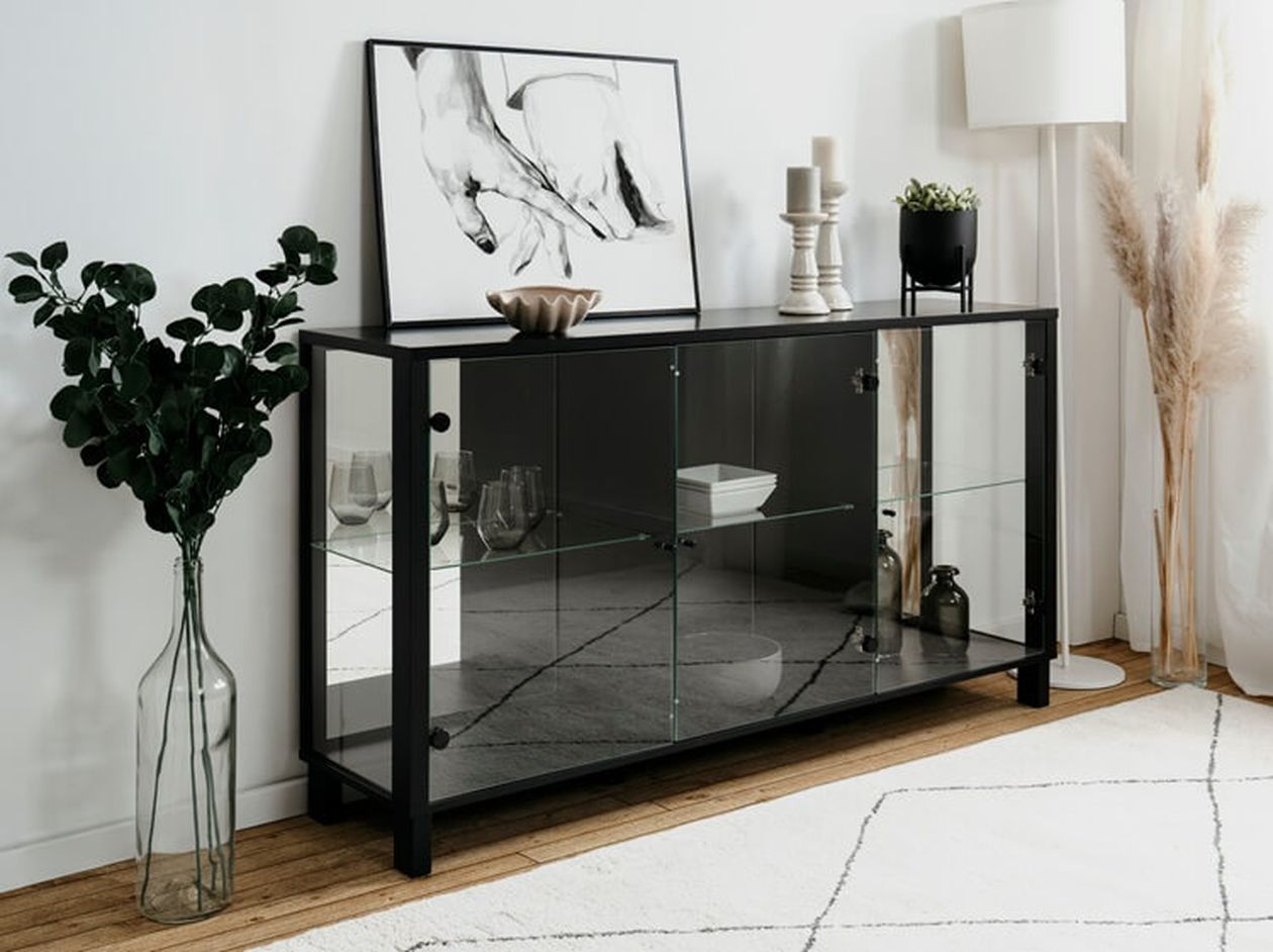 Schwarze Vitrine mit Glastüren und Glasböden in einem hellen Wohnzimmer, gefüllt mit Gläsern und Dekoobjekten, daneben Pampasgras und Eukalyptuszweige.
