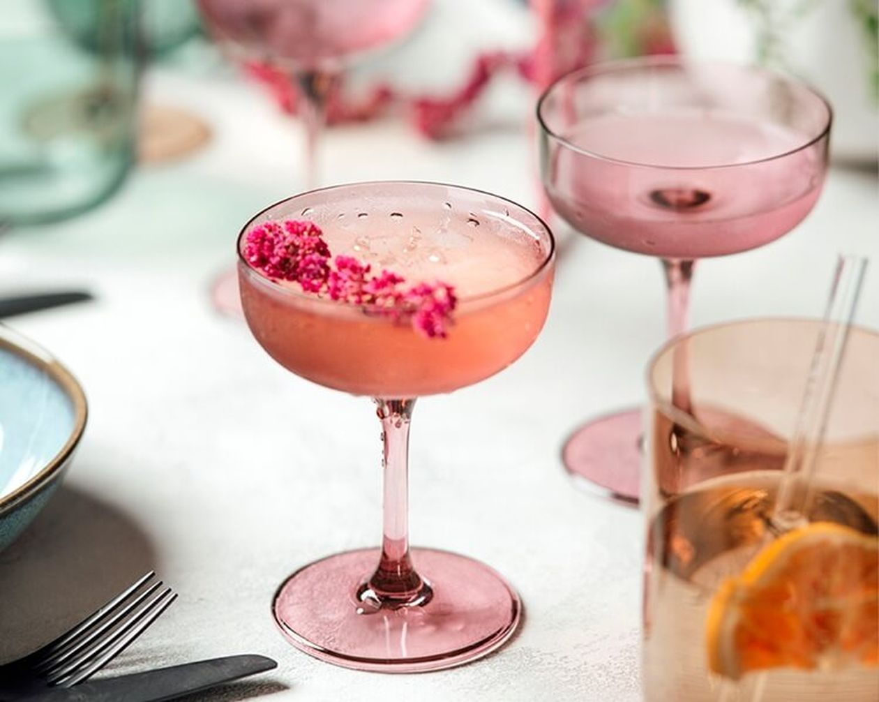Rosa Cocktail in einem Glas, garniert mit kleinen rosa Blüten, umgeben von ähnlichen Gläsern und Geschirr auf einer hellen Oberfläche.