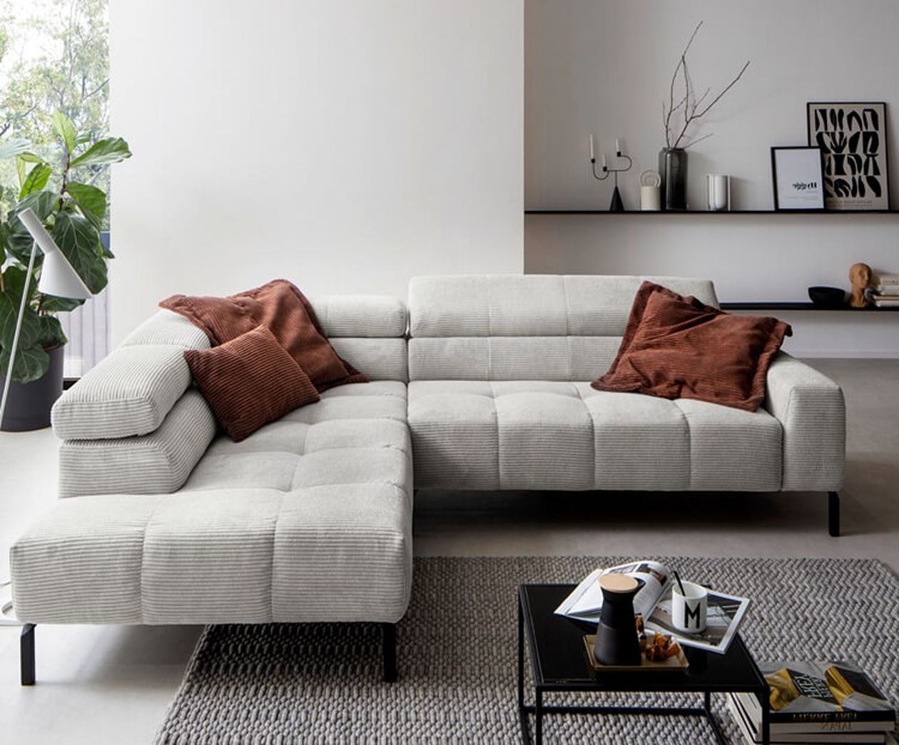 Hellgraues Cord-Ecksofa mit gesteppter Sitzfläche und rostfarbenen Kissen in einem skandinavisch eingerichteten Wohnzimmer mit schwarzem Metallregal.