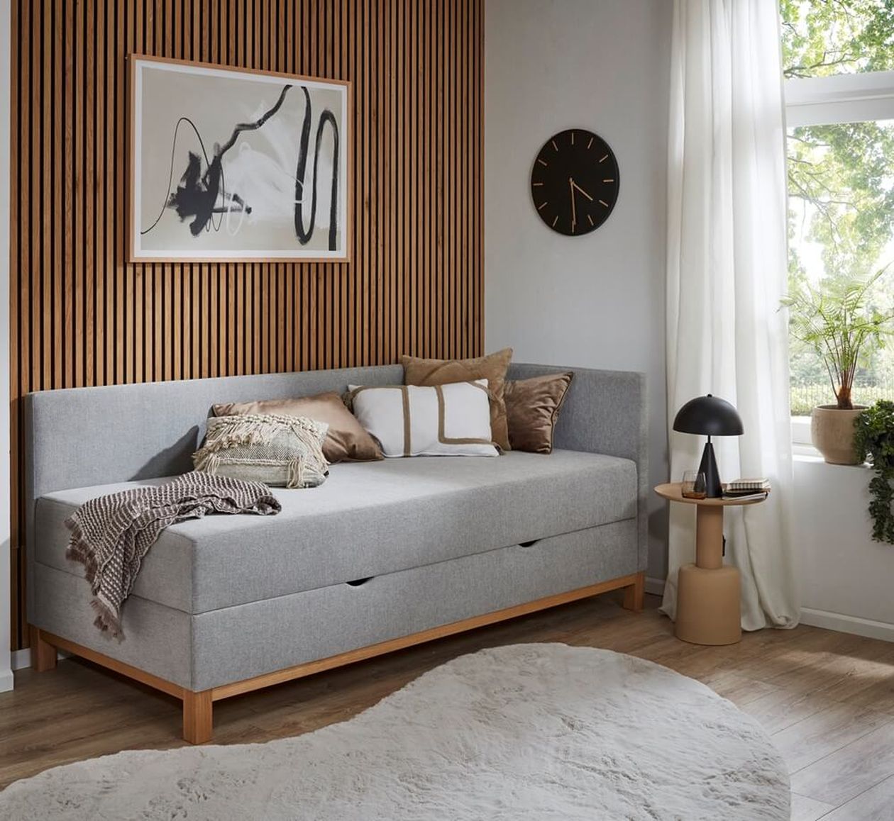 Ein gemütliches Zimmer mit einem grauen Tagesbett, abstrakter Kunst, einer Holzwand, einer runden Uhr und einem Fenster mit Pflanzen. Ein runder Teppich und ein kleiner Tisch mit einer Lampe runden den Look ab.