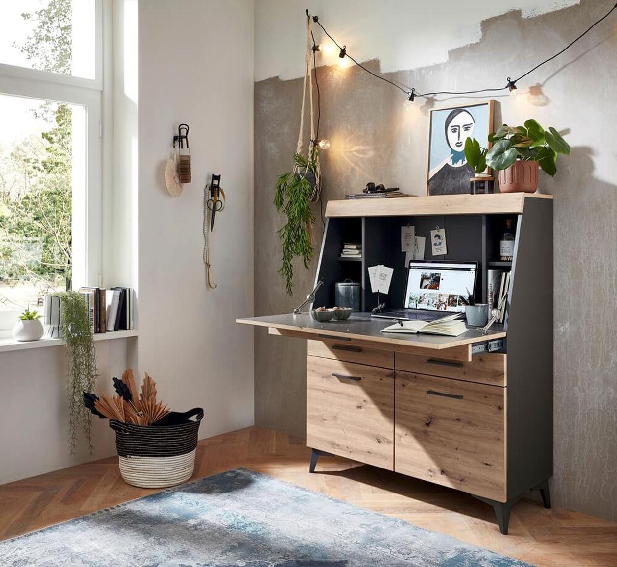 Gemütliche Home-Office-Ecke mit Holzschreibtisch, Laptop, Pflanzen, Wandkunst, Lichterketten und einem großen Fenster mit Blick ins Grüne.