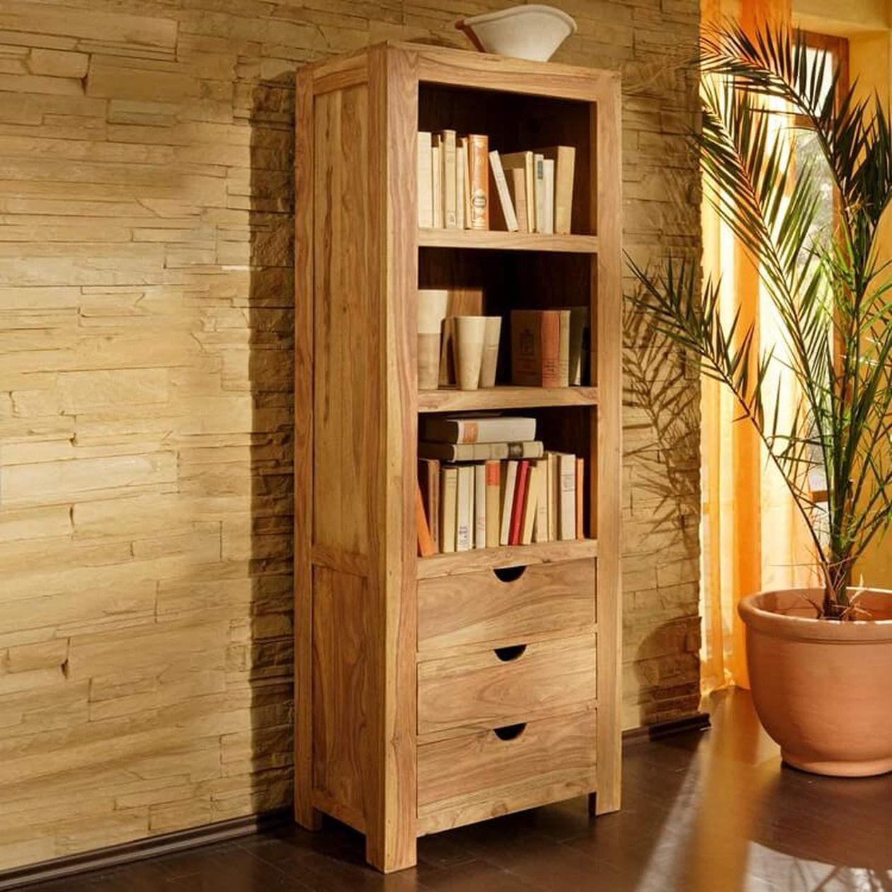 Hohes hölzernes Bücherregal mit Büchern und Dekoration, drei Schubladen unten, neben einer strukturierten Steinwand und einer großen Topfpflanze.