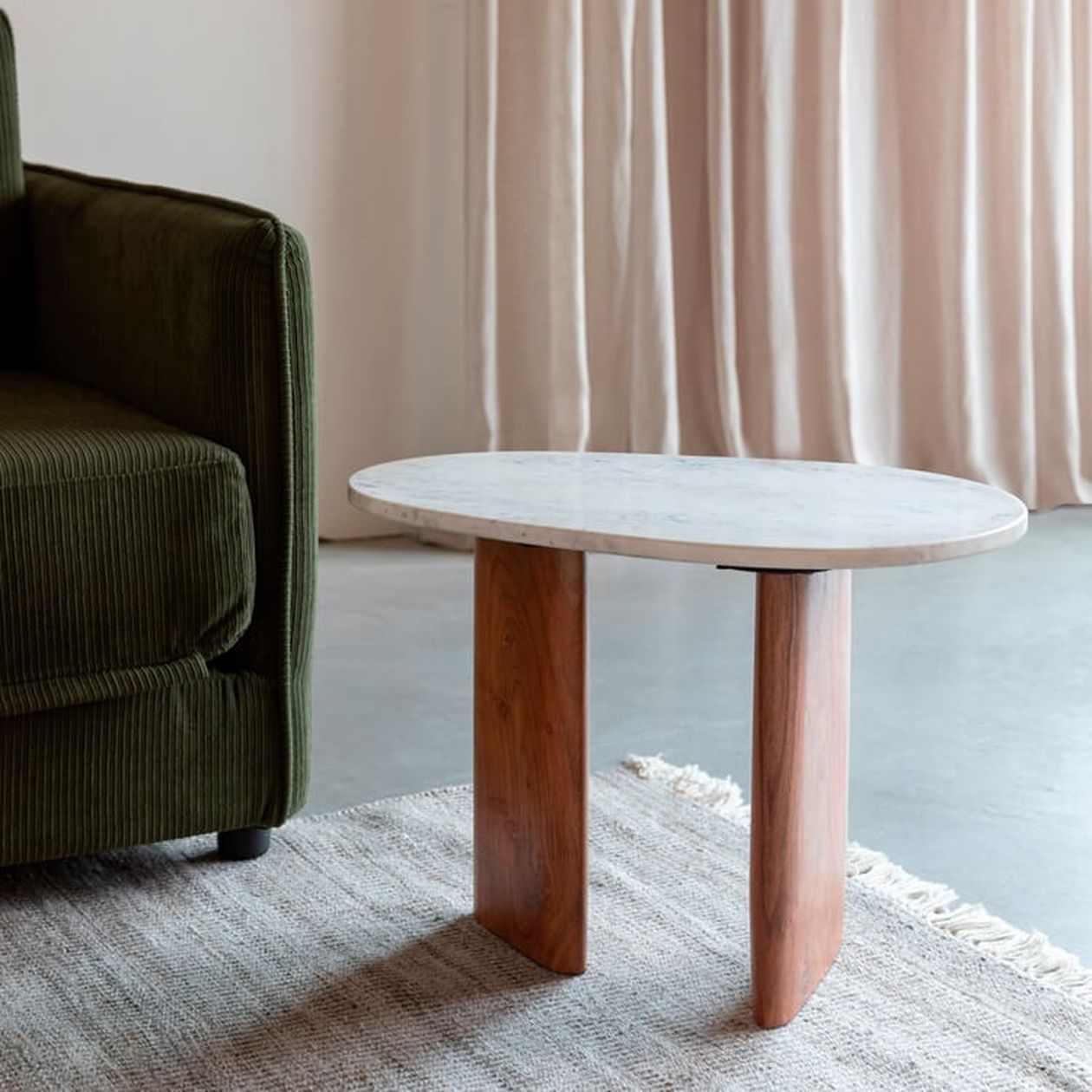 Ovaler Beistelltisch mit weißer Marmorplatte und zwei massiven Holzbeinen, neben einem grünen Cord-Sessel auf hellem Teppich.