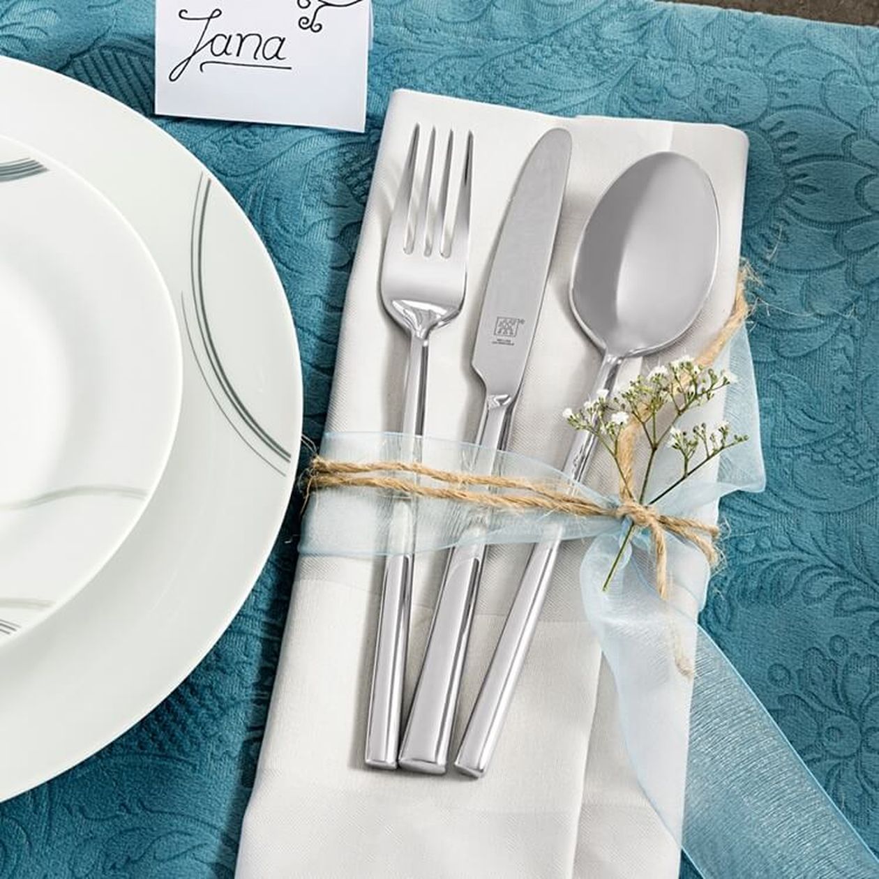 Silbernes Besteck (Gabel, Messer, Löffel) mit Juteschnur und Schleierkraut auf einer weißen Stoffserviette, daneben ein weißer Teller auf türkisfarbenem Tischtuch mit Platzkarte „Jana".