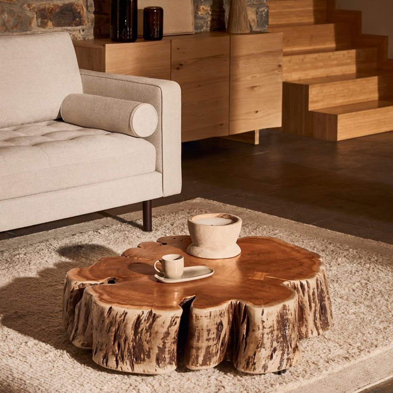 Niedriger Couchtisch aus einer massiven Baumscheibe mit natürlicher Rindenstruktur, darauf Keramikschalen, in einem warmen Wohnzimmer mit Holztreppe.