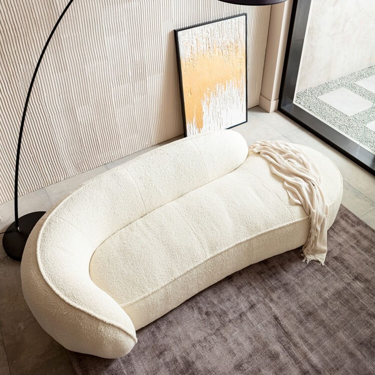 Geschwungene weiße Bouclé-Recamiere mit organischer Form auf grauem Teppich, daneben eine Bogenlampe und ein abstraktes Wandbild.