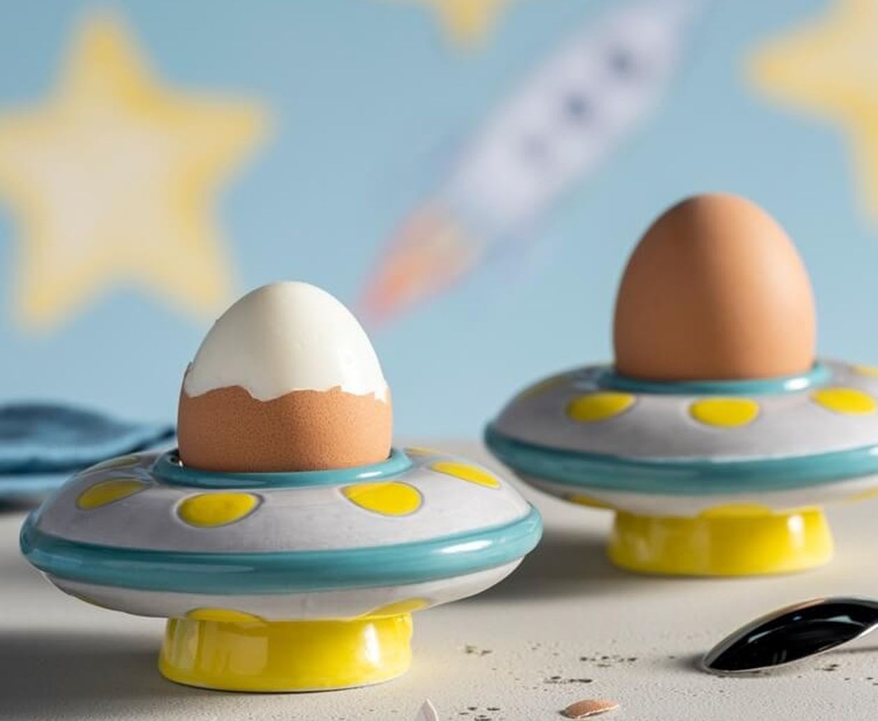 Zwei keramische Eierbecher in UFO-Form mit türkis-gelben Punkten, jeweils mit einem Frühstücksei, vor blauem Hintergrund mit gelben Sternen und einer Rakete.